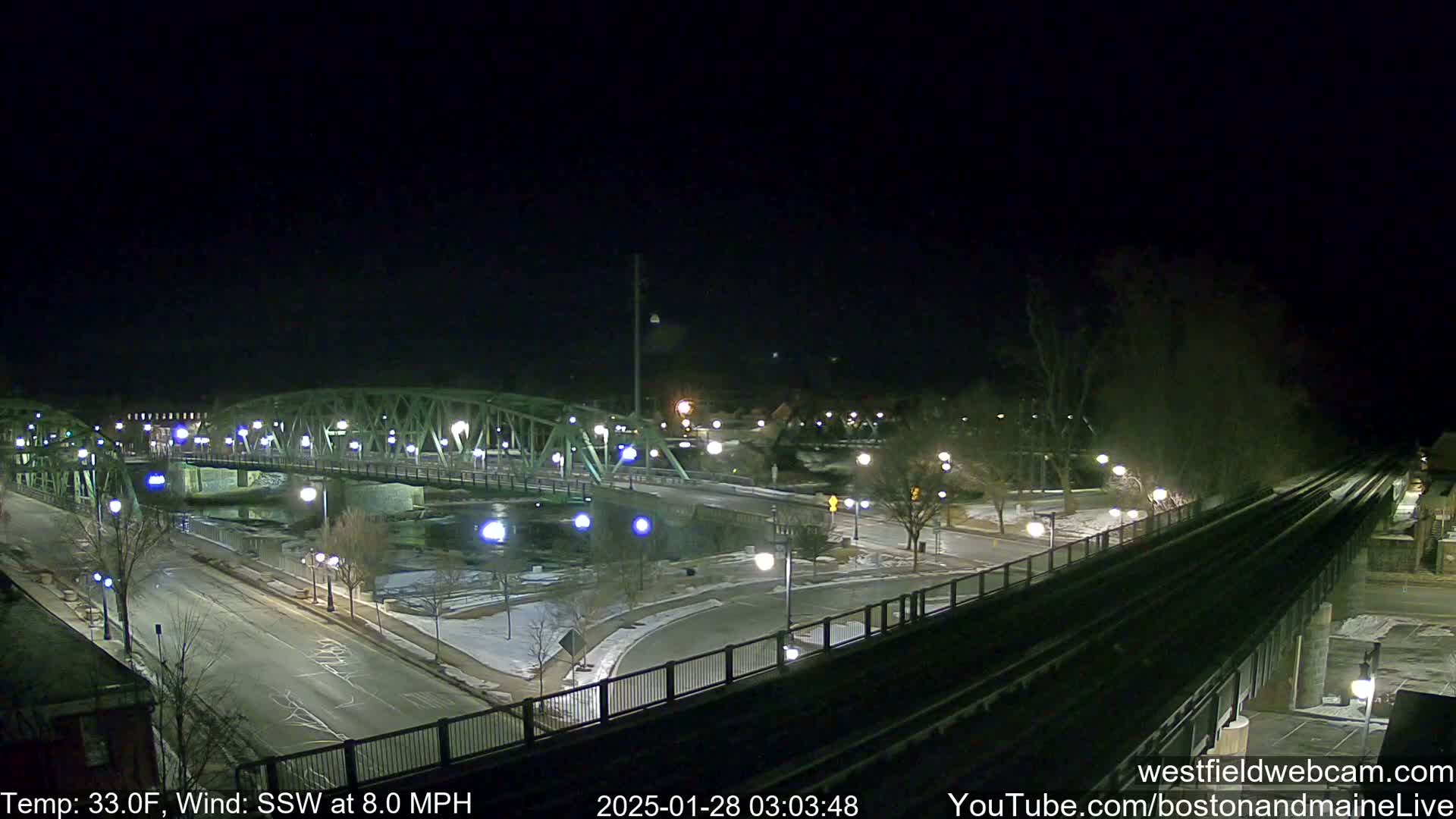 A nighttime view of a lit-up bridge over a river, with a roadway and train tracks visible, under a clear, dark sky, with a temperature of 33°F and a south-southwest wind at 8.0 mph.