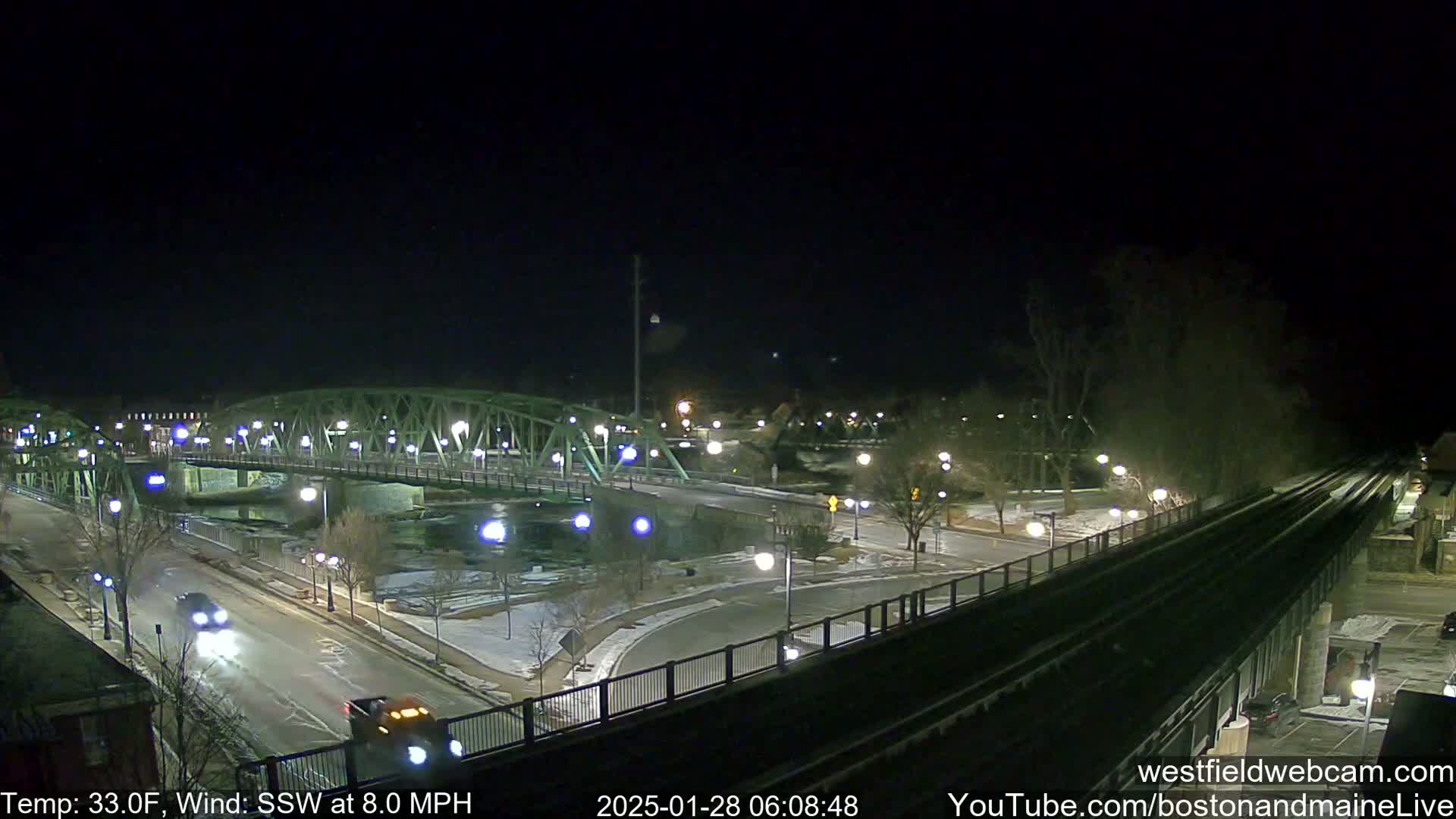A nighttime view of a bridge over a river, with sparsely trafficked roads and lightly snow-covered ground under a clear, dark sky; the temperature is 33°F, and the wind is blowing from the south-southwest at 8 mph.