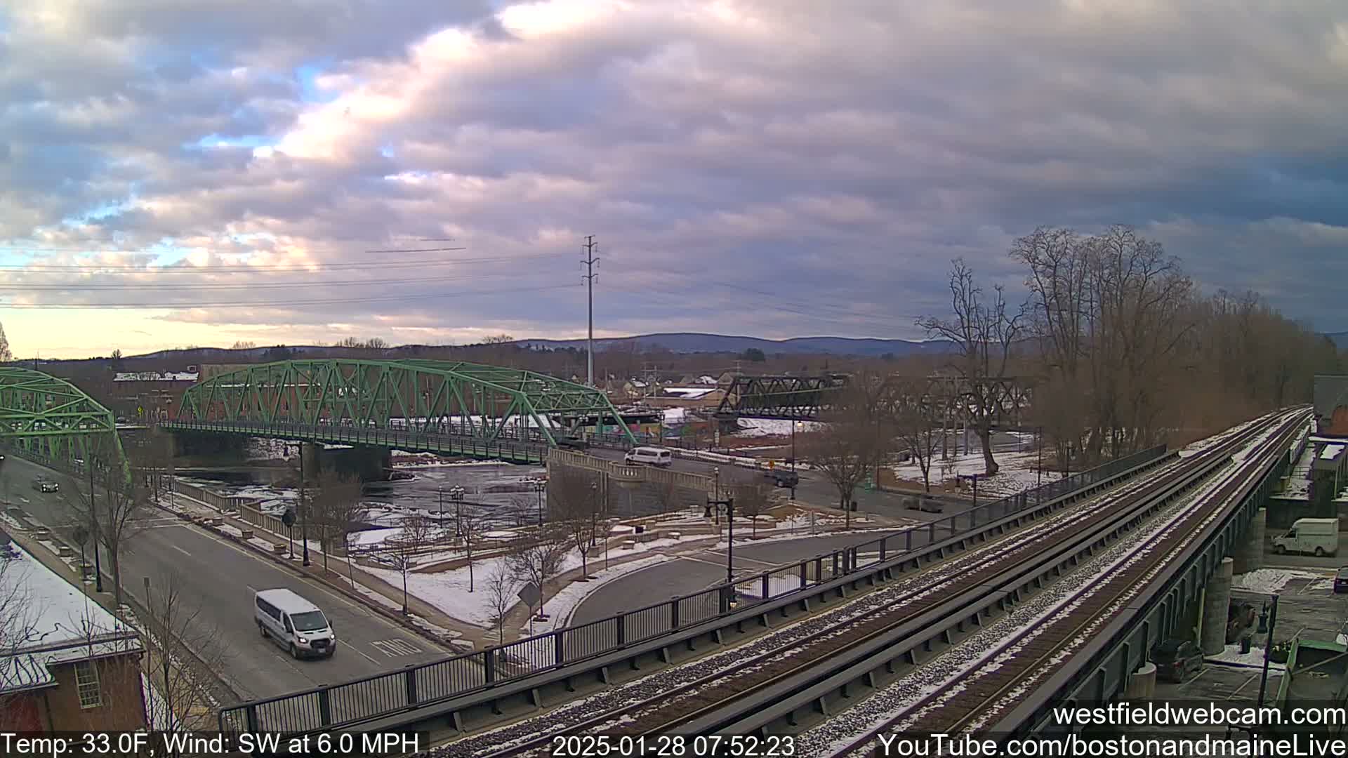 A partly cloudy day shows a green bridge over a river with train tracks and a road intersecting near it, all lightly dusted with snow.