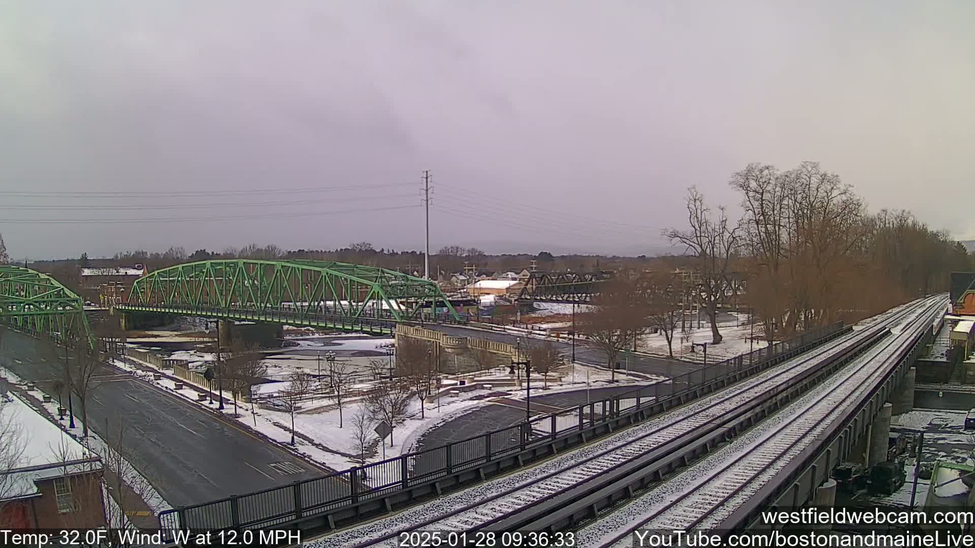 A snow-covered scene shows two green bridges spanning a river, with train tracks in the foreground under an overcast sky.