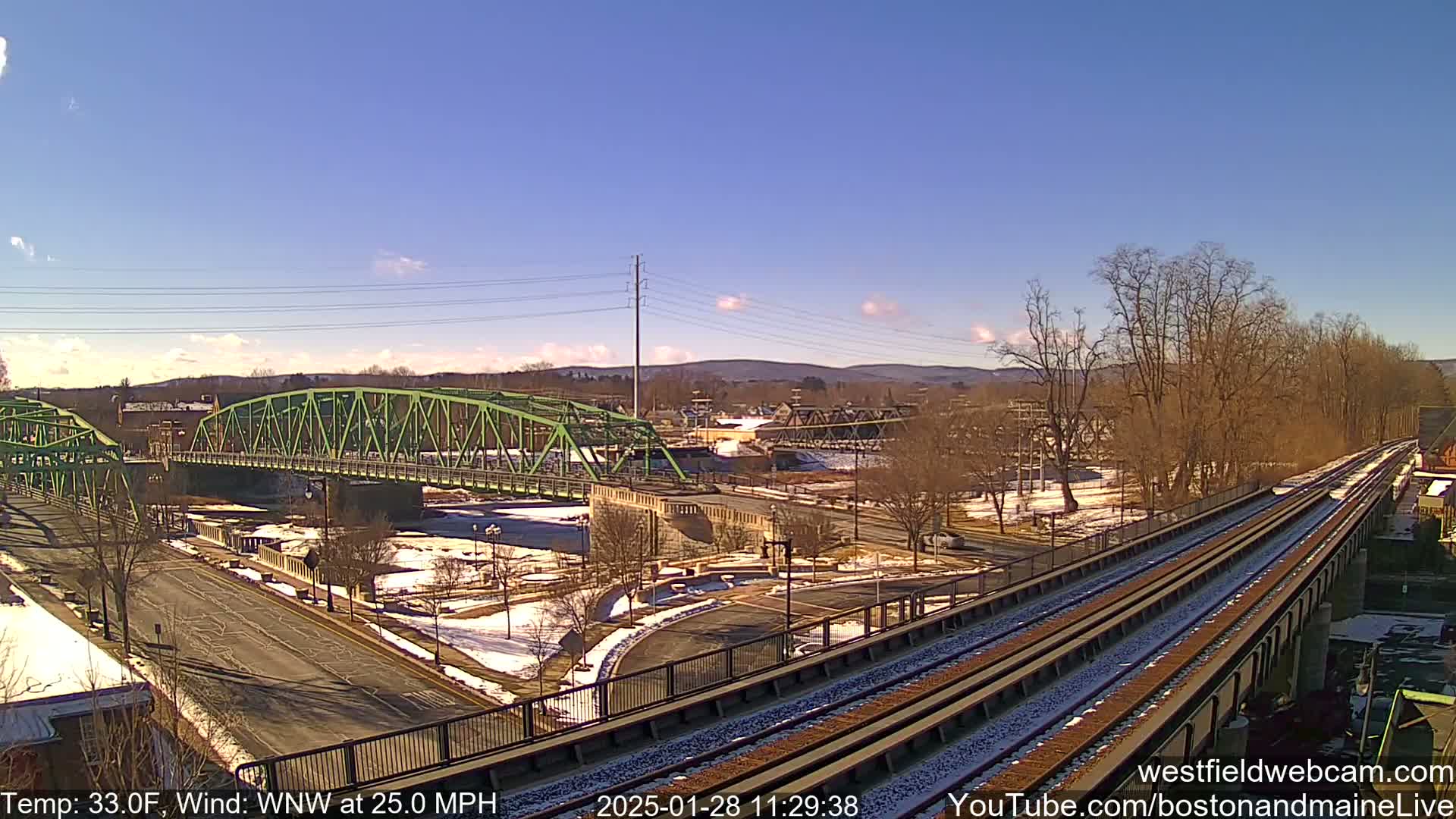 A sunny day with a strong wind shows a view of train tracks and a green bridge over a river, with some snow on the ground and bare trees along the banks.