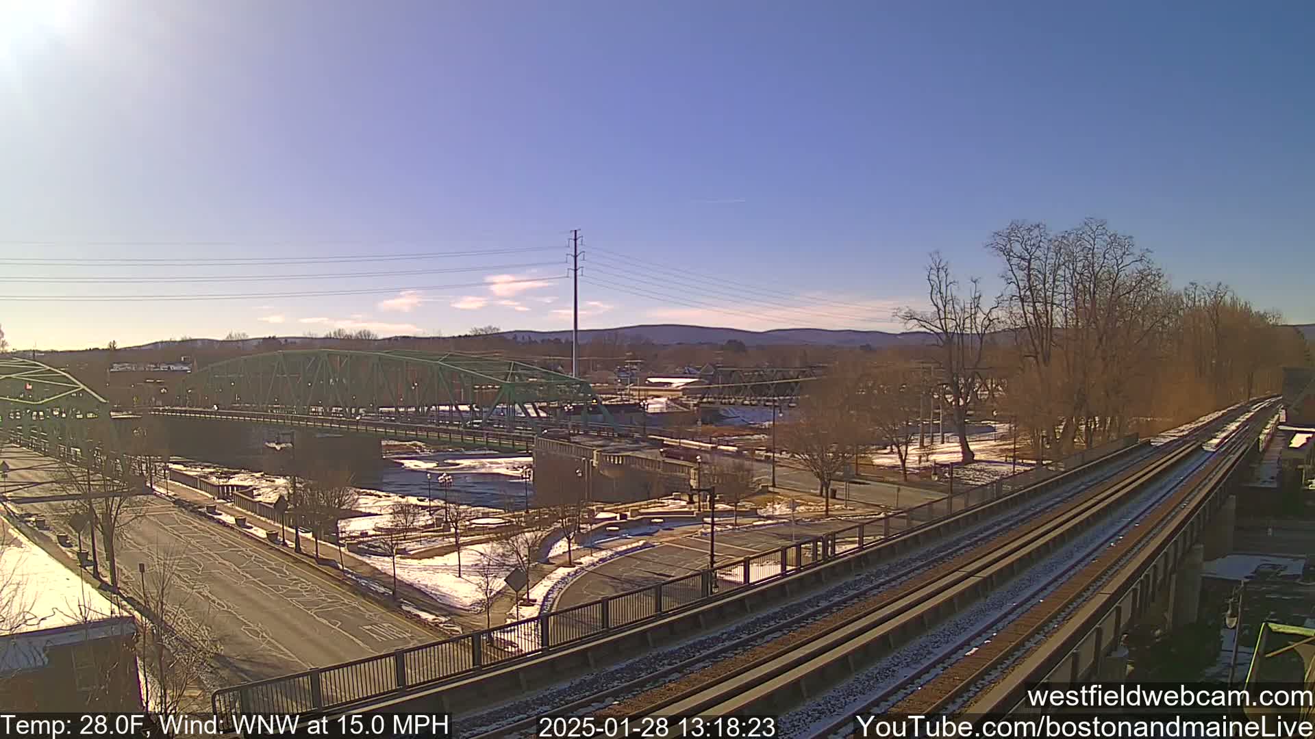 A view of a river with two bridges and a train track, under a clear blue sky with a light dusting of snow on the ground.