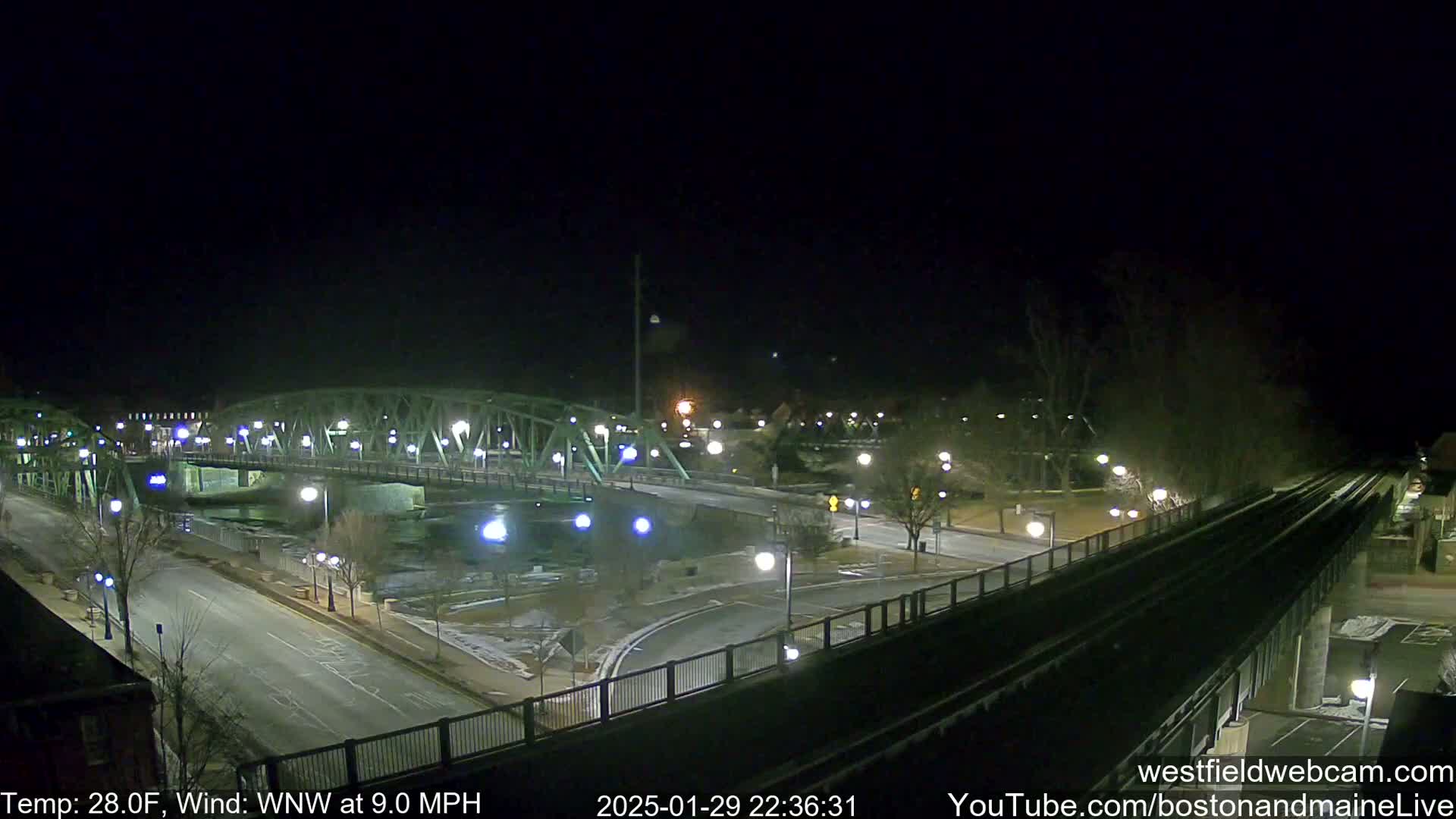 A nighttime view of a bridge over a river, with roadways and streetlights illuminating a sparsely populated scene under a clear, dark sky, and a temperature of 28 degrees Fahrenheit with a wind from the west-northwest at 9 miles per hour.