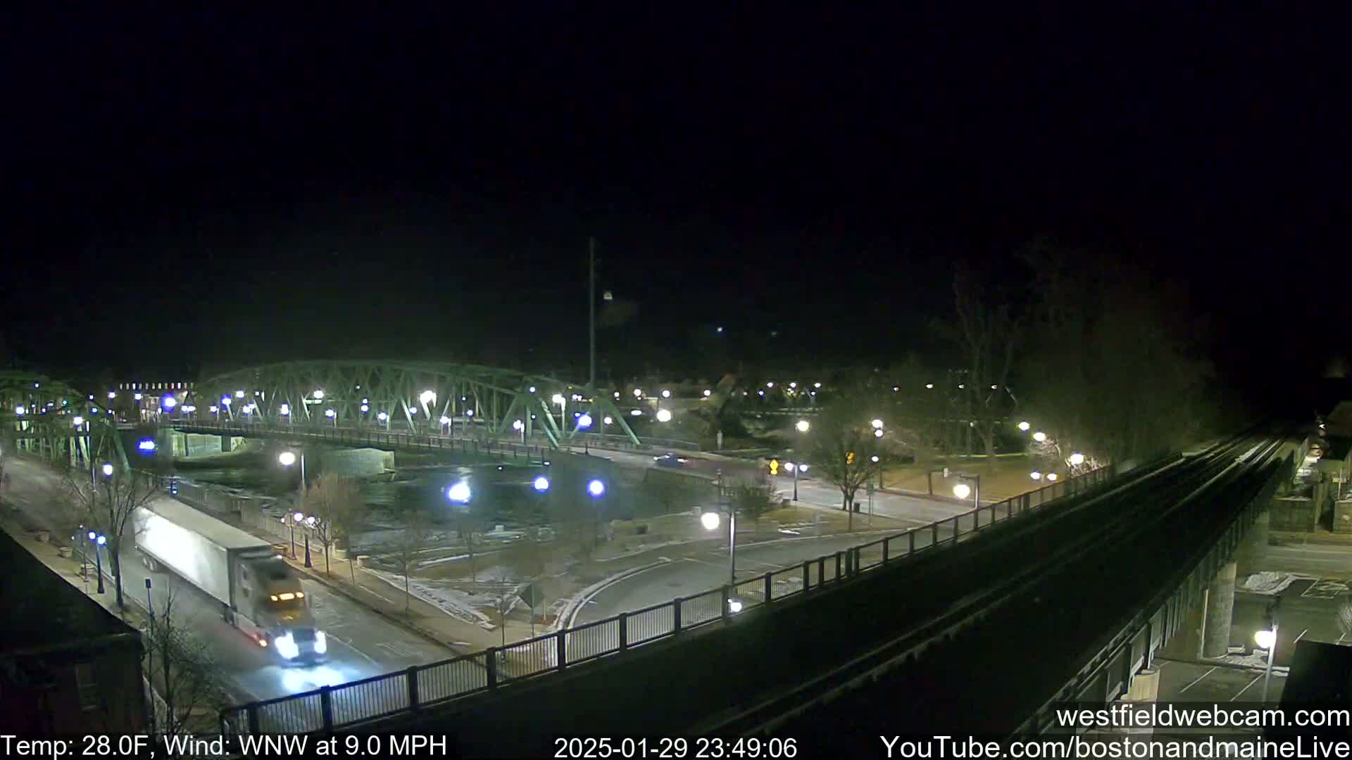 A nighttime, high-angle view shows a bridge over a river, a roadway with a semi-truck, and train tracks, all under a clear, cold sky with a wind blowing from the west-northwest.