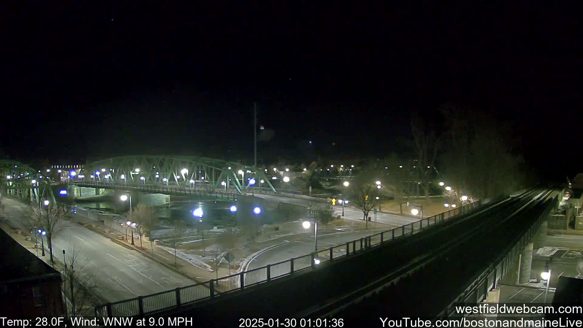 A nighttime view of a green metal bridge over a dark river, with a roadway and train tracks intersecting below, under a clear, dark sky and a 28°F temperature with a 9.0 mph wind from the west-northwest.