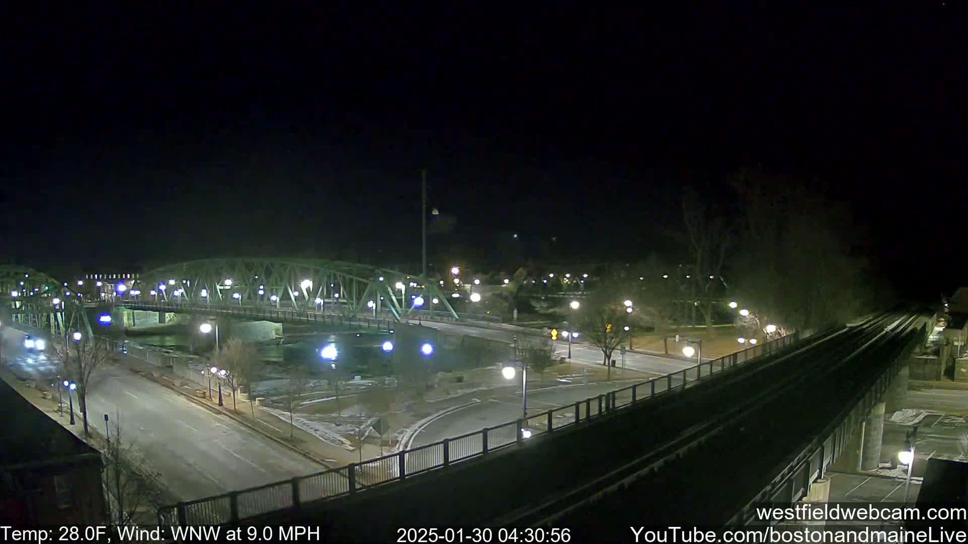 A nighttime view of a bridge over a river, with a roadway and train tracks visible, under a clear, cold sky with a wind from the west-northwest at 9.0 mph.