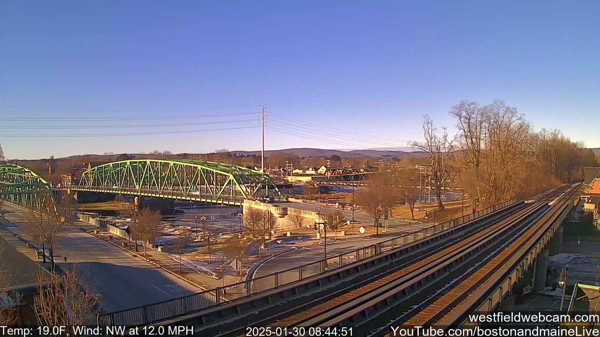 A green bridge spans a river, with train tracks and a town visible in the background under a clear, sunny sky.