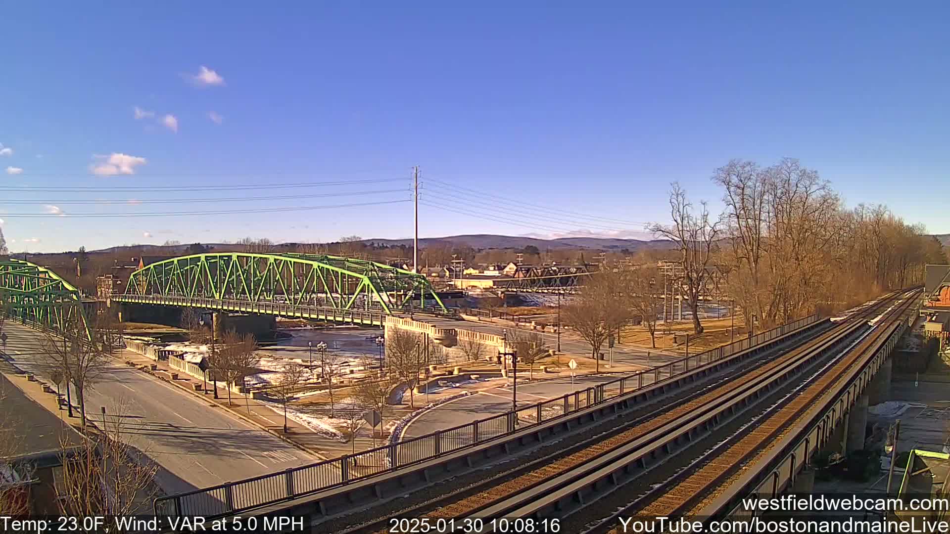 A bright, sunny day shows a view of a green bridge over a river, a roadway, and train tracks leading toward a town nestled in a valley under a clear blue sky.