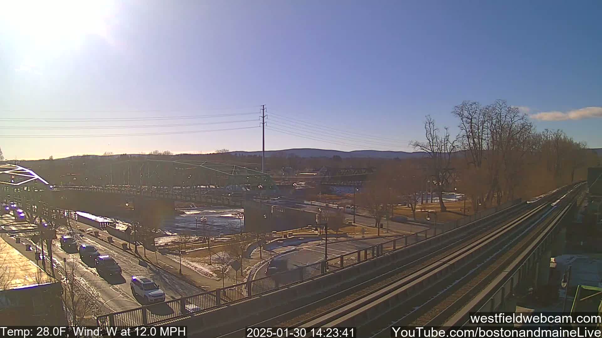 A sunny day with a west wind at 12 mph shows a view of a bridge over a river, a roadway with cars, a train track, and some bare trees.