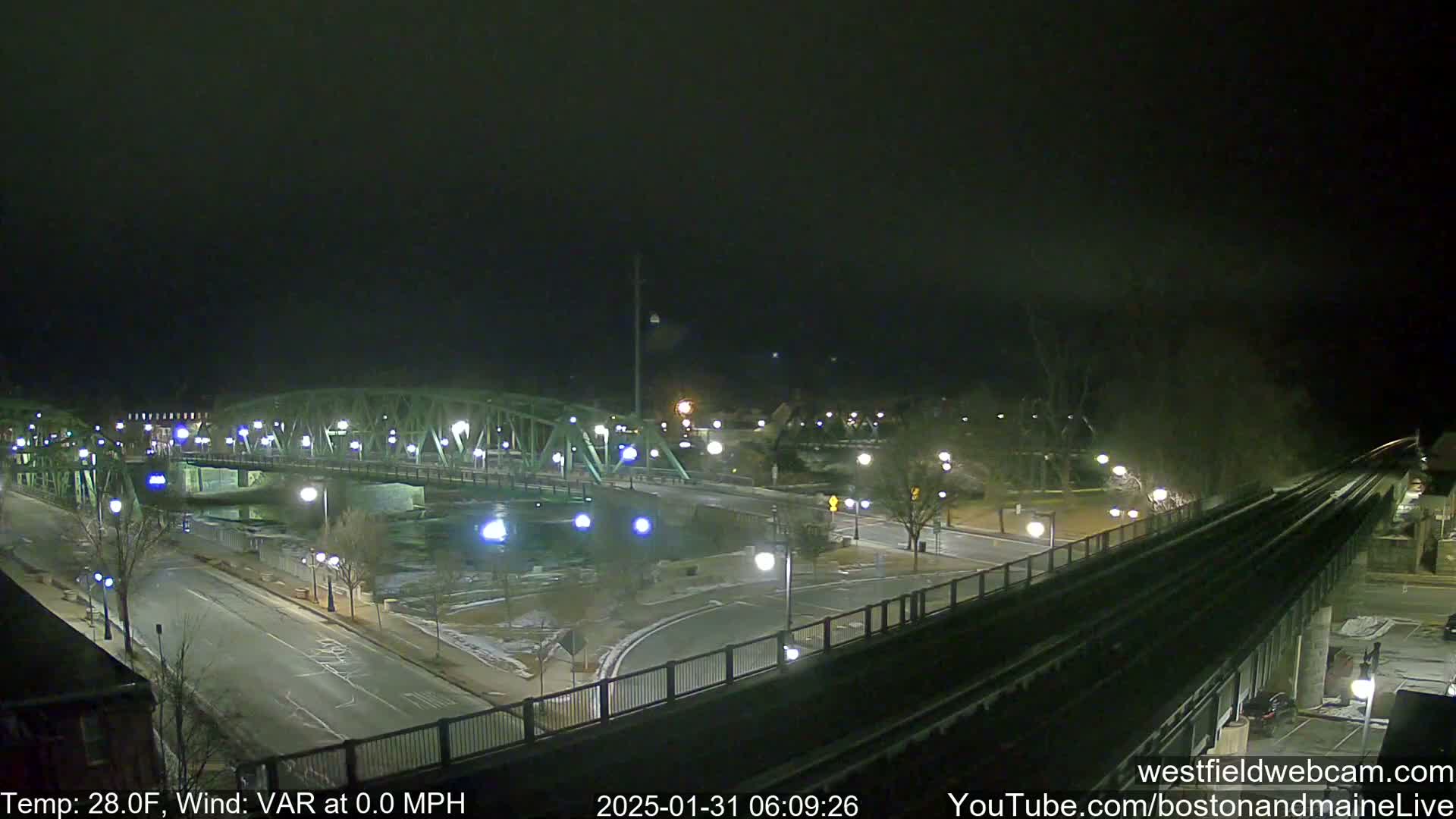 A nighttime view of a bridge over a river, illuminated streets, and train tracks, with a temperature of 28 degrees Fahrenheit and calm winds.