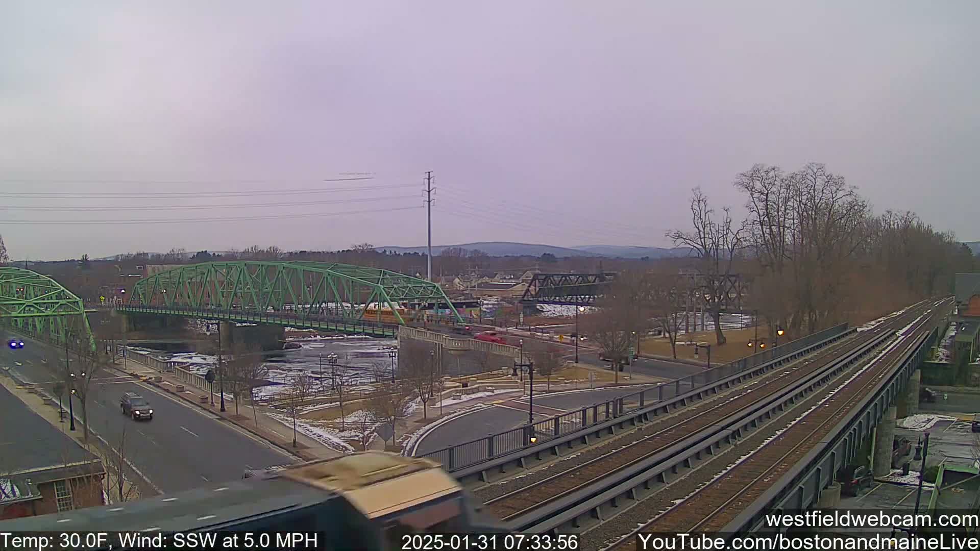 A view of a town with two bridges over a river, train tracks, and a road, under an overcast sky.