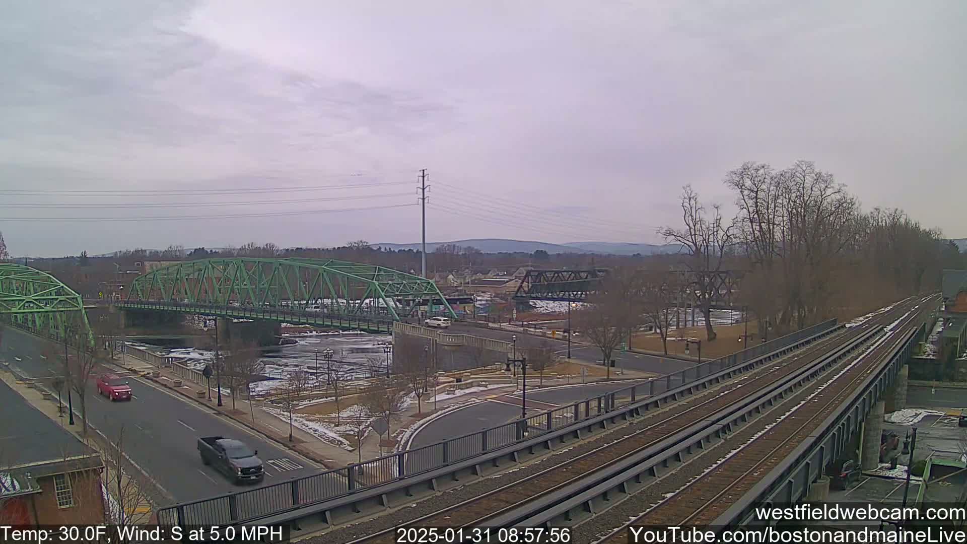 A view of a town with two bridges spanning a river, some cars on roads, and train tracks elevated above a roadway, all under a cloudy sky.