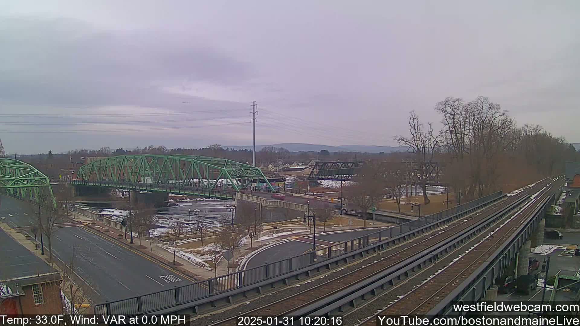 A view of two bridges over a river, with a railroad track in the foreground, under an overcast sky with a temperature of 33°F and a wind speed of 0.0 MPH.