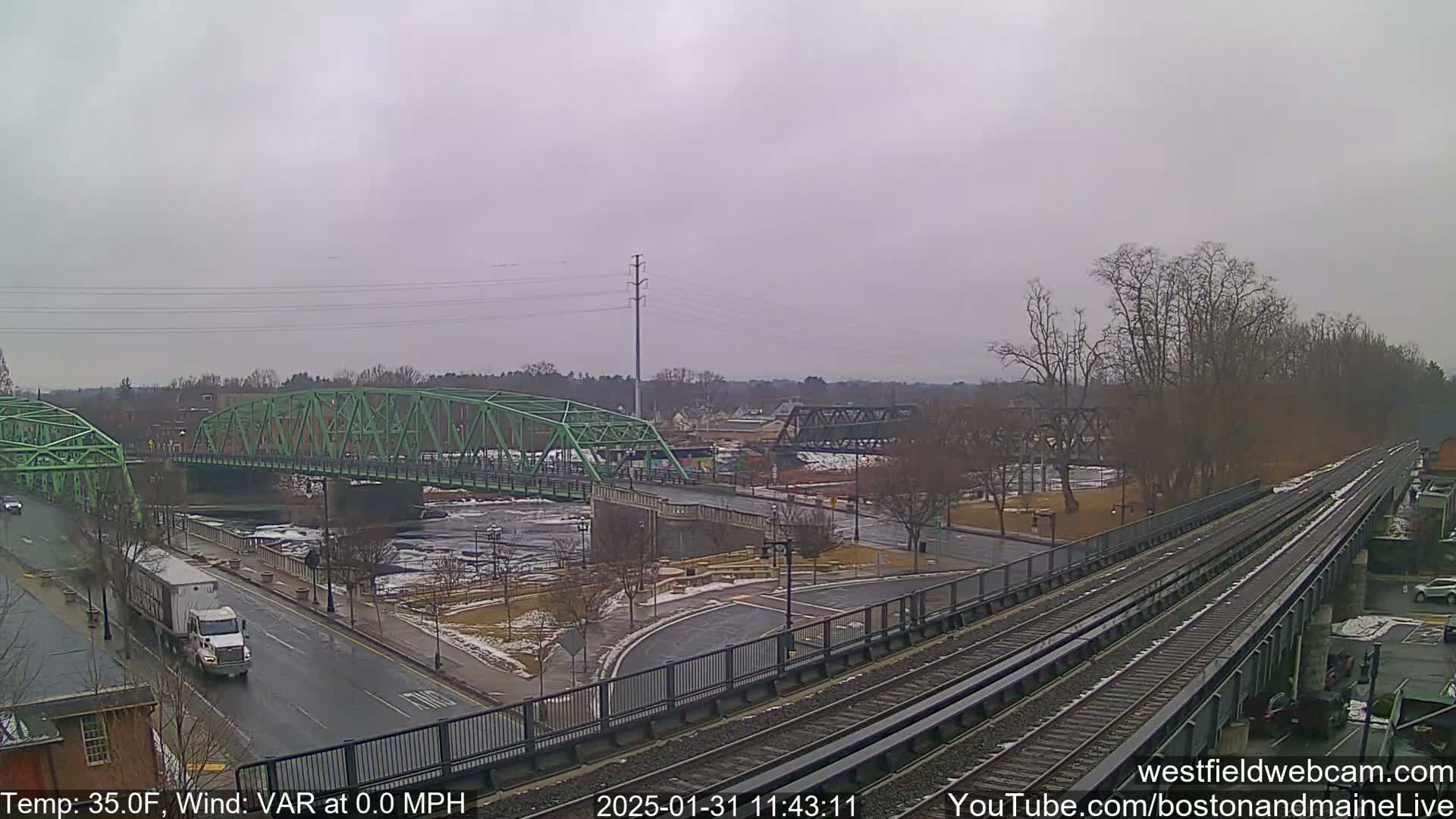 A view of a green bridge over a river, with a train track beside it, on an overcast day with a temperature of 35 degrees Fahrenheit and no wind.