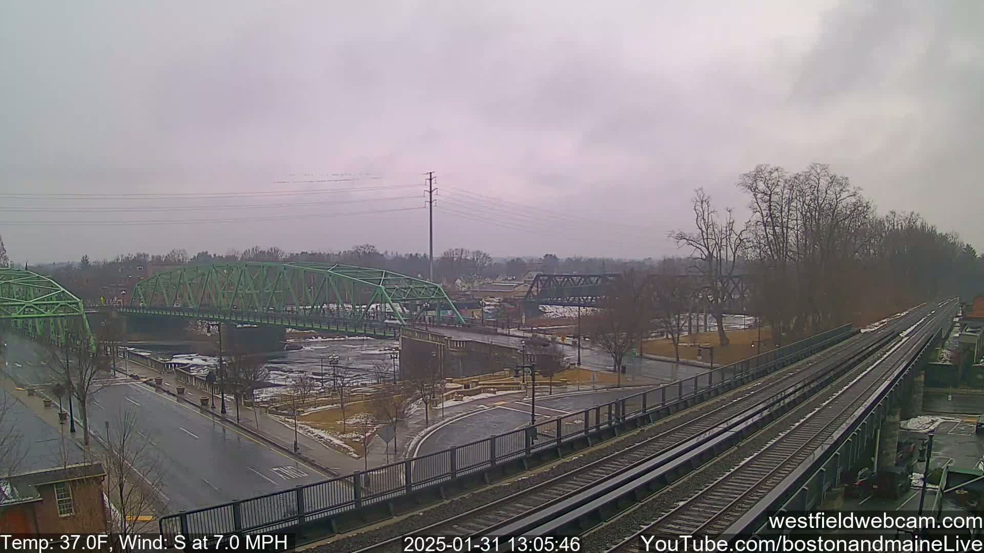 A view of two bridges over a river, train tracks, and a road intersection under an overcast sky.