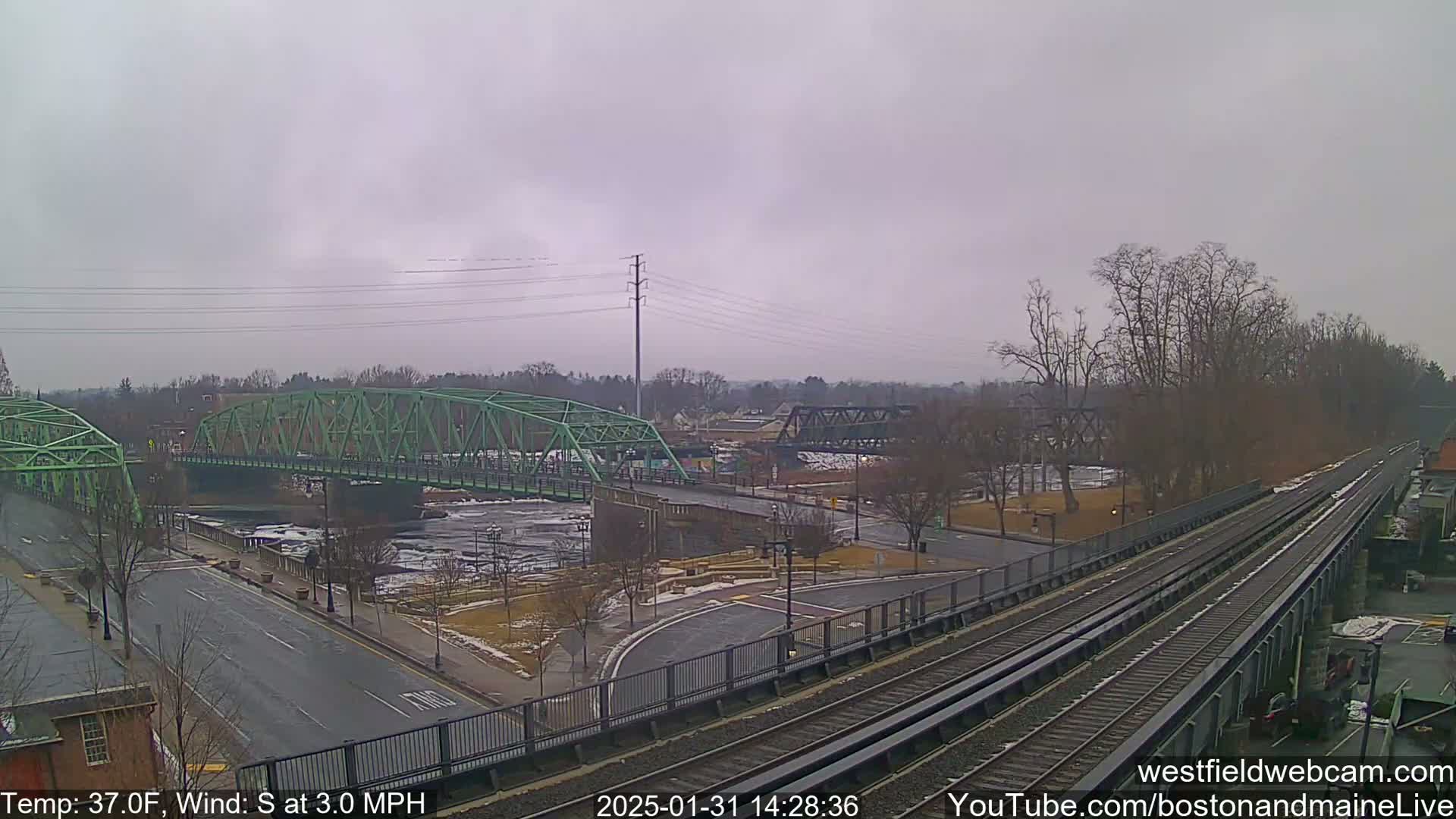 A view of two bridges over a river, with train tracks and roadways intersecting, under an overcast sky with a south wind.