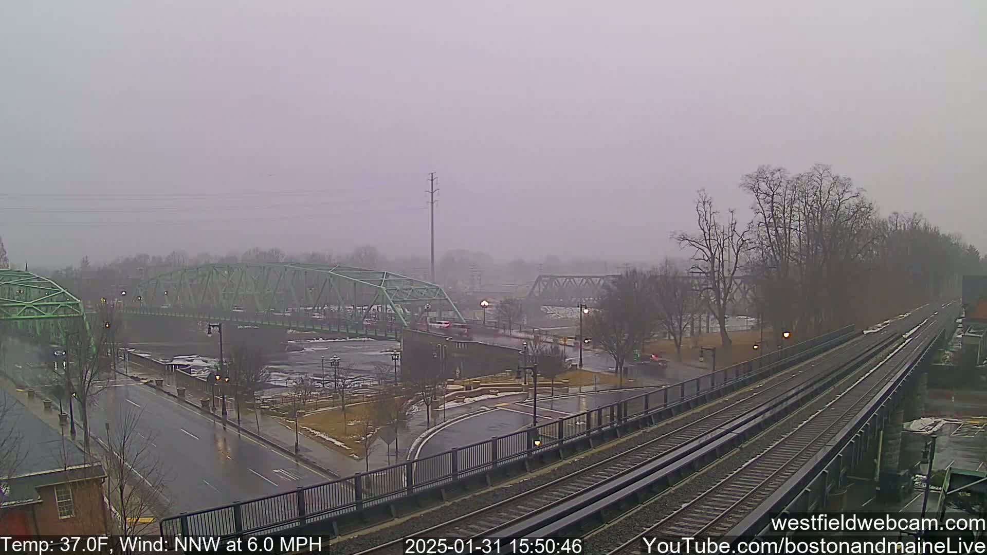 A rain-swept view shows a river, two bridges, train tracks, and roadways, with bare trees in the background under a gray sky.