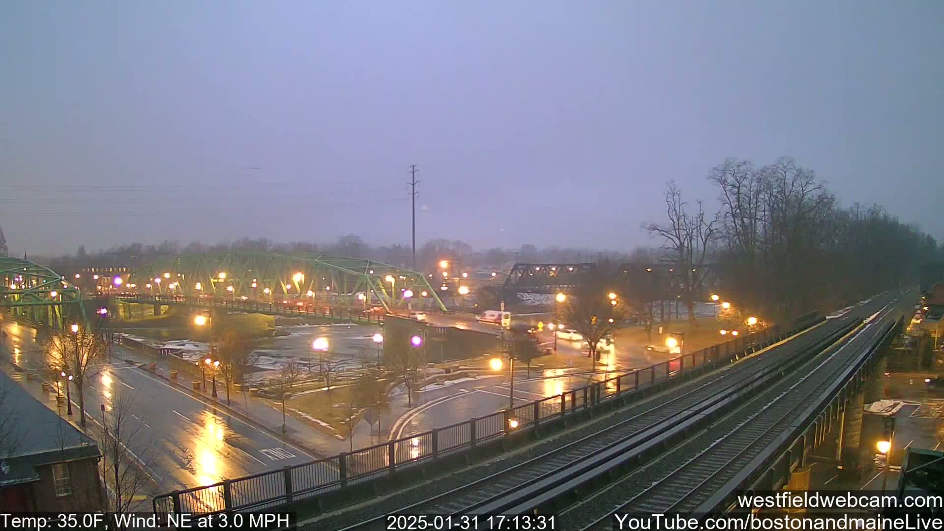 A wet, nighttime scene shows a roadway intersecting with train tracks near two bridges, one green and one dark-colored, under a slightly overcast sky with a northeast wind.
