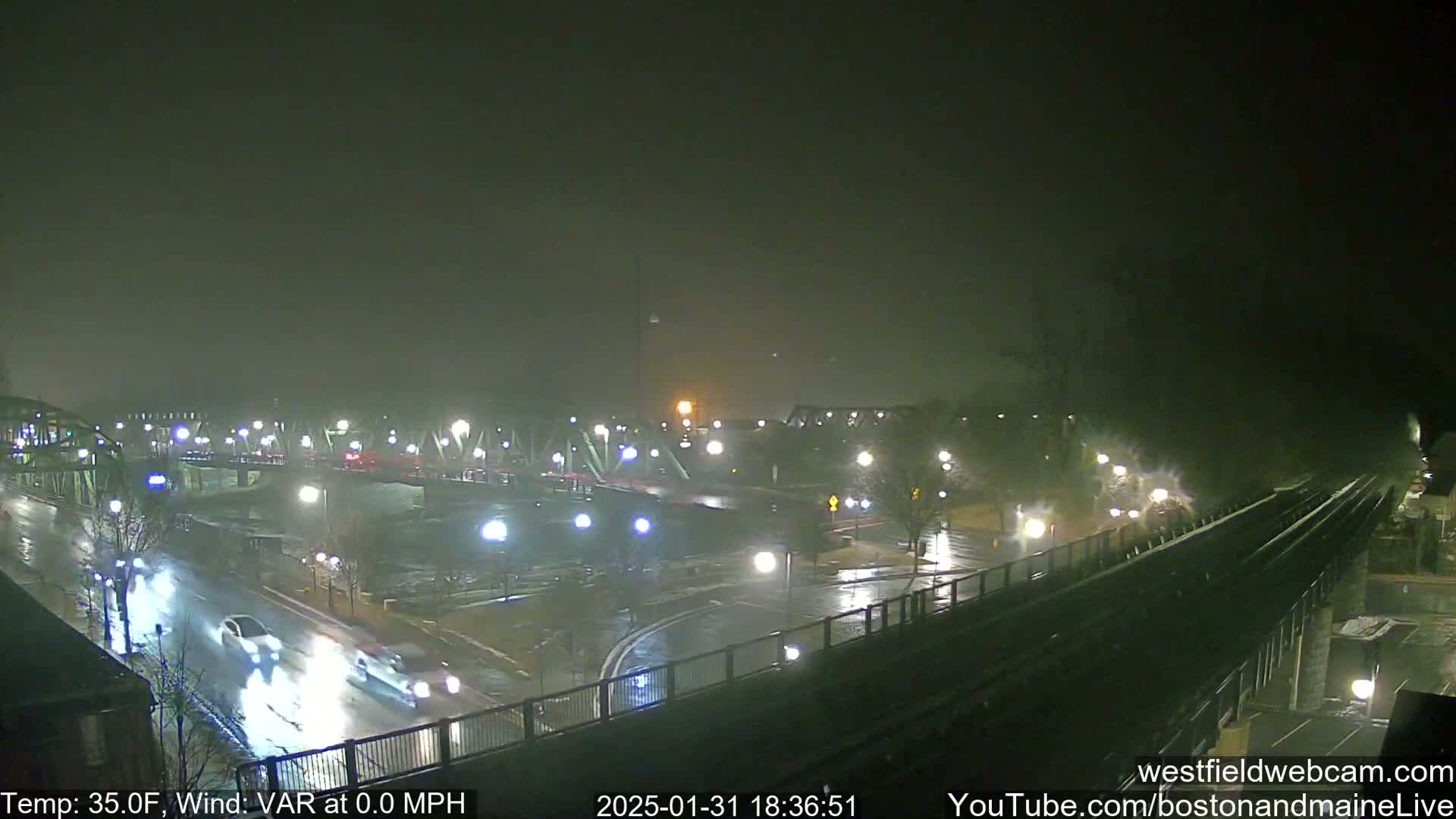 A rain-slicked nighttime street scene shows a bridge, cars, and streetlights under a dark, overcast sky.