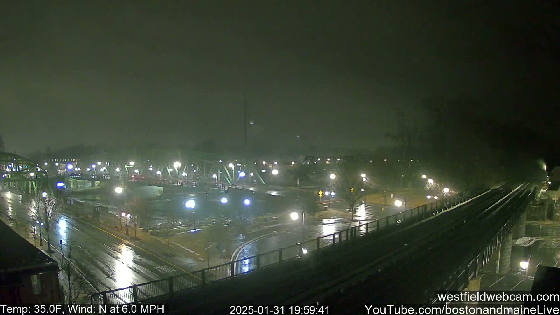 A rain-slicked, nighttime cityscape with a bridge and train tracks, under a dark, overcast sky.