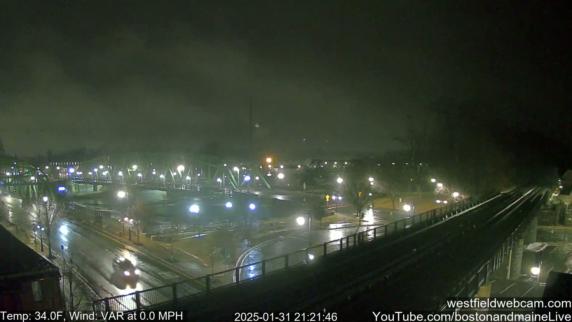 A rain-slicked nighttime cityscape view shows a bridge, roads, and a railway track under a dark, cloudy sky.