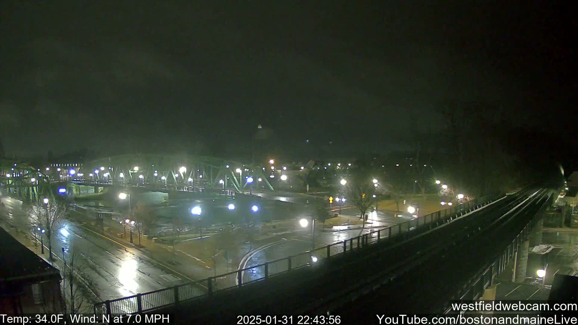 A nighttime view of a wet city street scene with a bridge and train tracks, under a dark, overcast sky.
