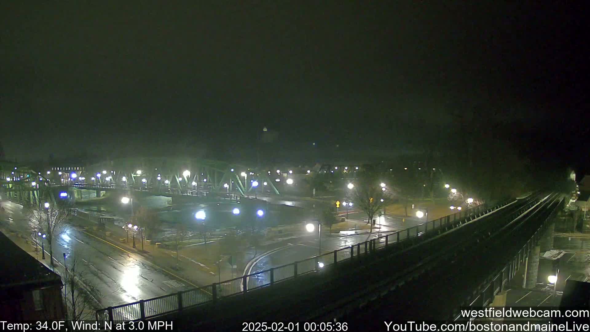 A wet, nighttime scene shows a bridge and roads with streetlights reflecting on the wet pavement under a dark, overcast sky.