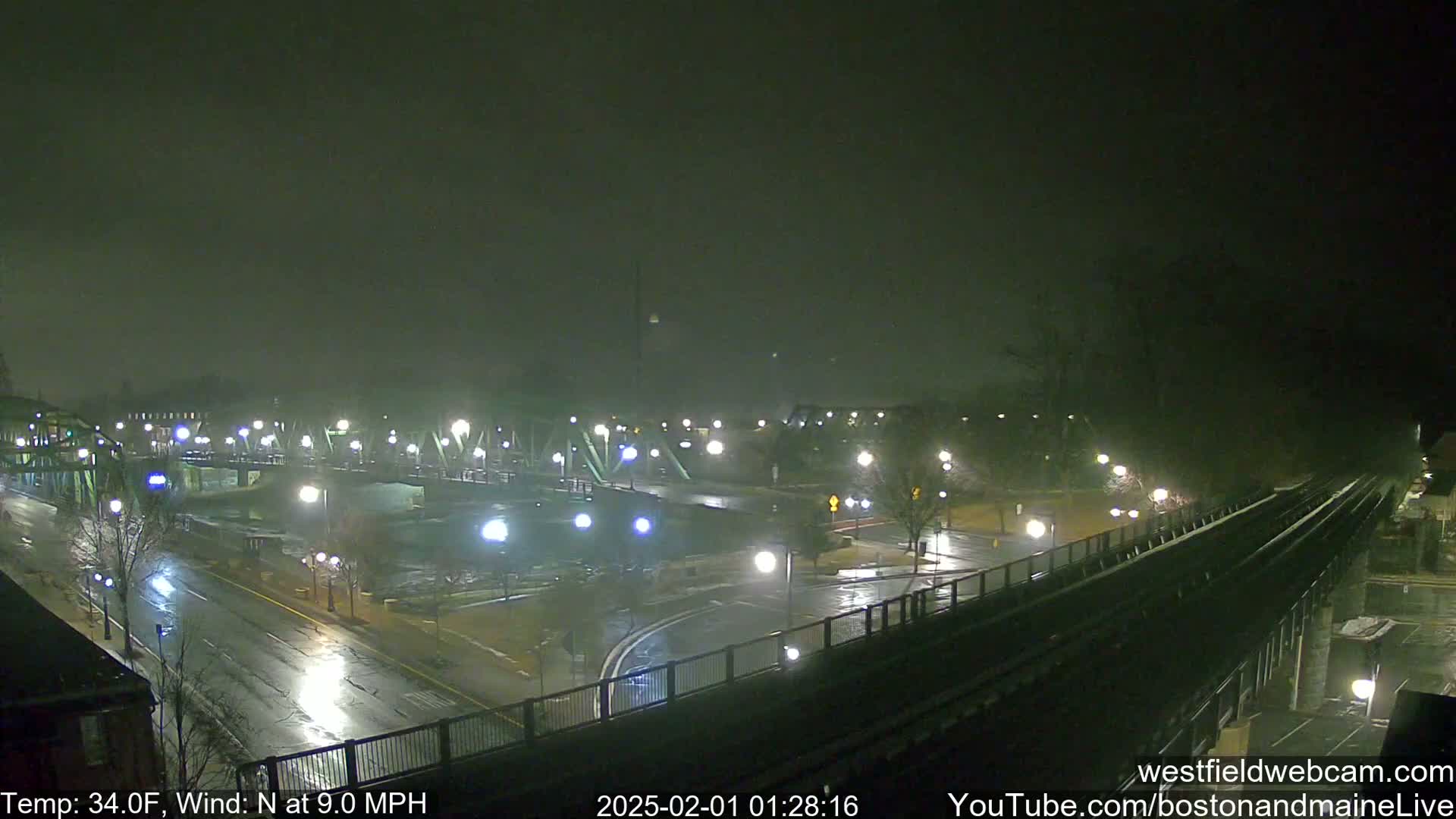 A nighttime view of a wet, dimly lit city street and bridge under foggy conditions, with a railway elevated above.