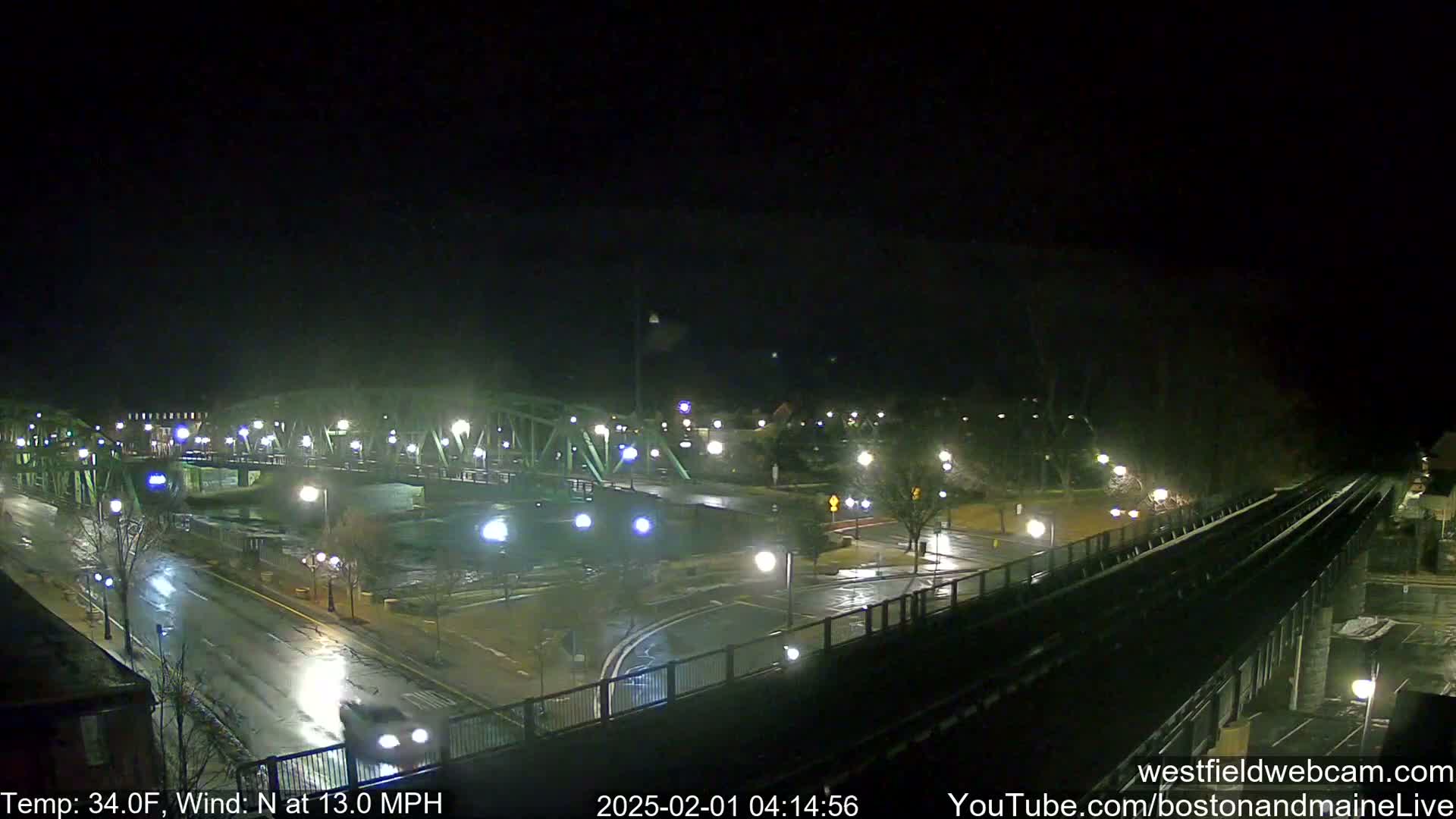 A wet, nighttime view of a bridge and roadways, with a train track in the foreground, under a dark sky with a north wind.