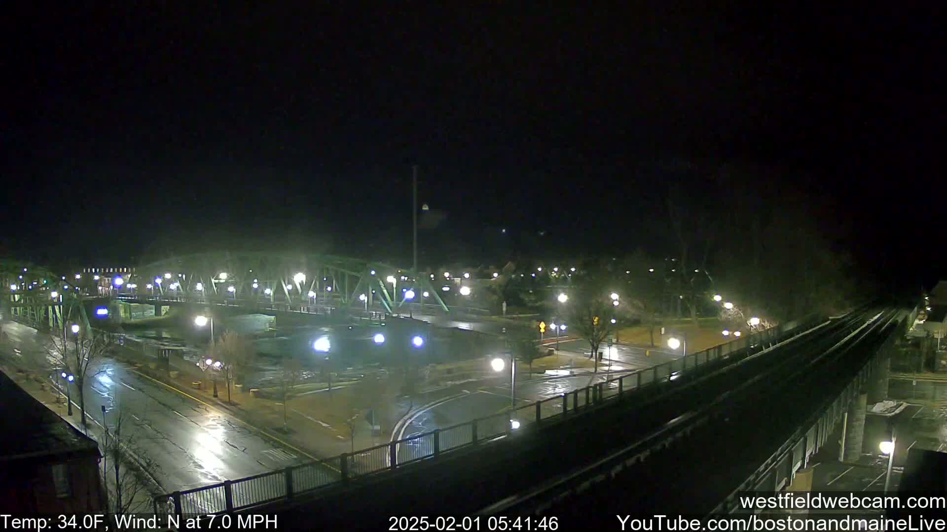 A rain-slicked nighttime street scene shows a bridge, a roadway intersection, and train tracks, with streetlights illuminating the wet surfaces under a dark sky.