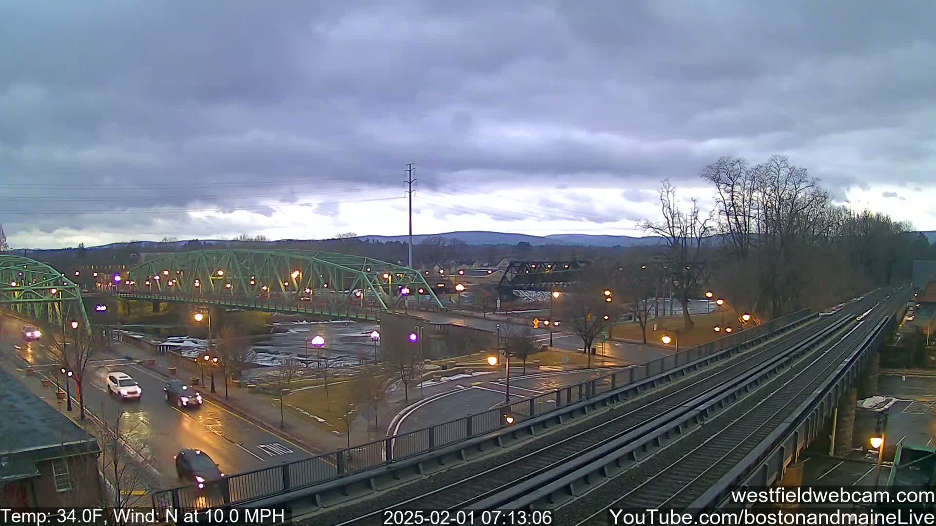 A view of a town under a cloudy sky shows a green bridge over a river, a roadway with cars, and a set of train tracks.