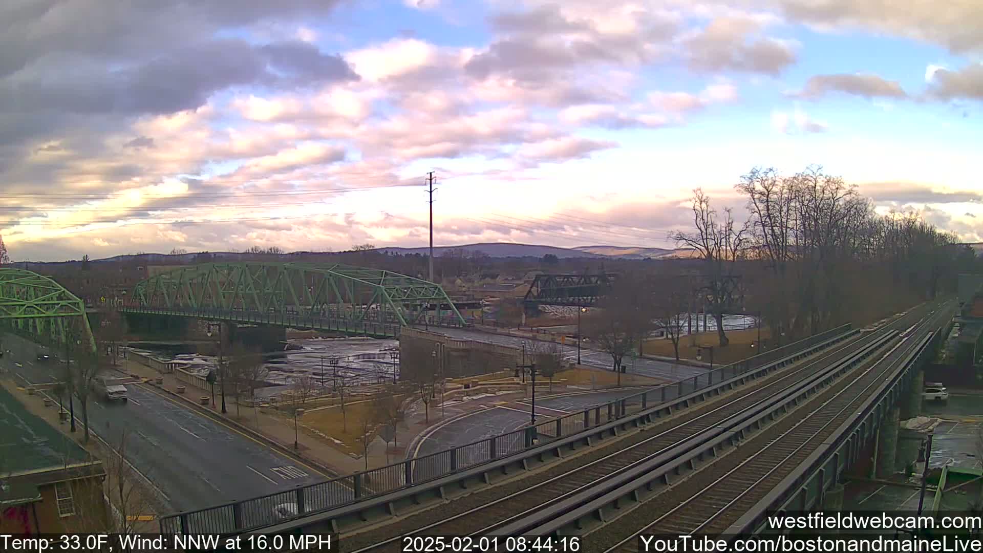 A partly cloudy, cool day reveals a view of two bridges over a river, with train tracks and a roadway intersecting in the foreground.