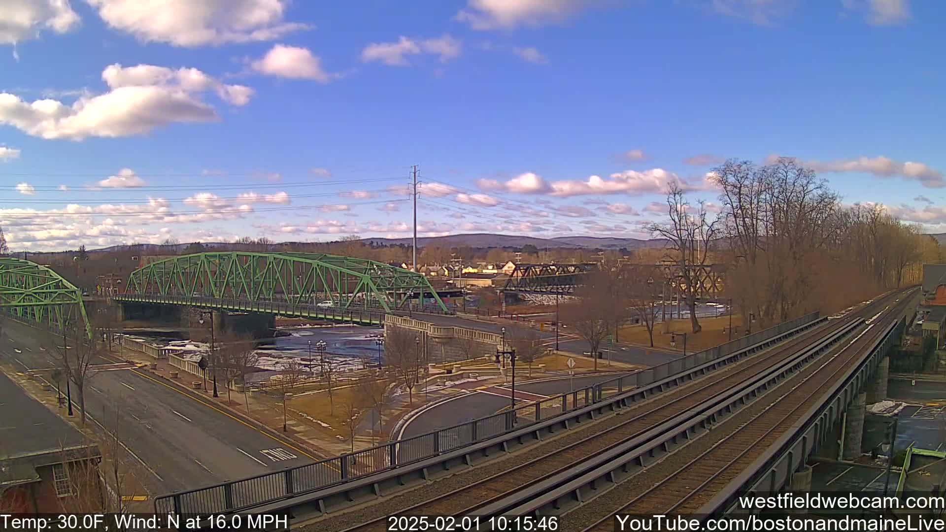 A partly cloudy day with a north wind at 16 mph shows a view of two bridges over a river, with train tracks in the foreground.