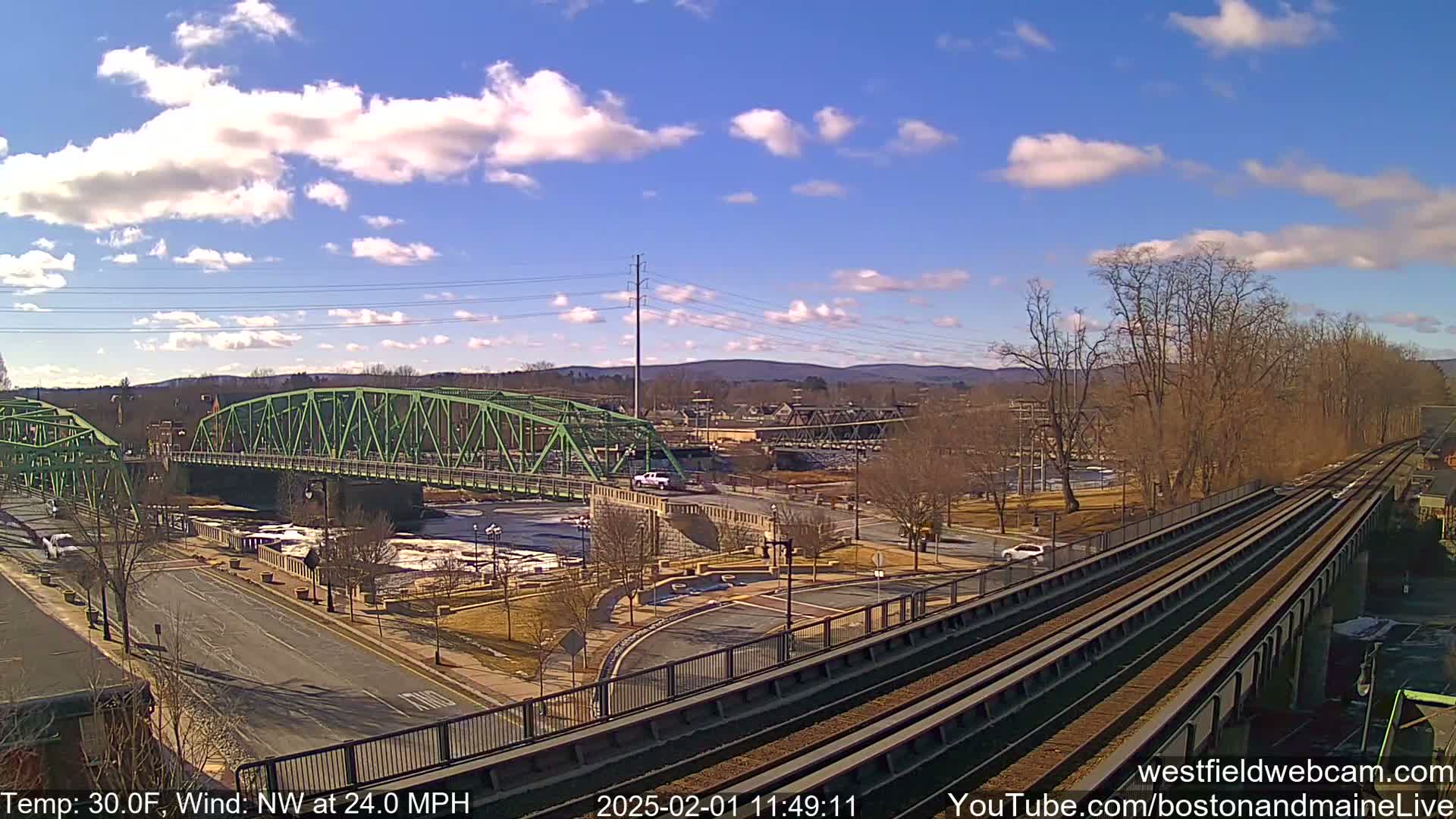 A view of a town with a green bridge over a river, under a partly cloudy sky with a strong northwest wind.