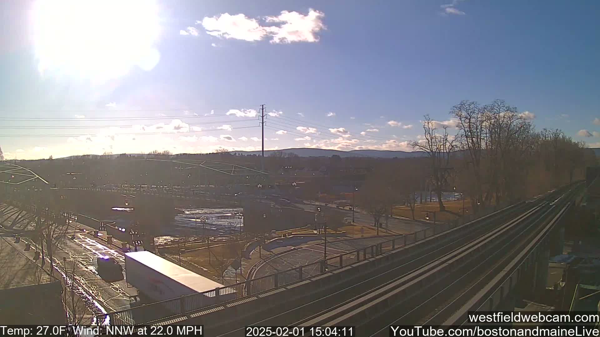 A sunny day with a north-northwest wind at 22 mph shows a view of a roadway, a parking lot, and train tracks from an elevated vantage point, with bare trees and distant hills in the background.