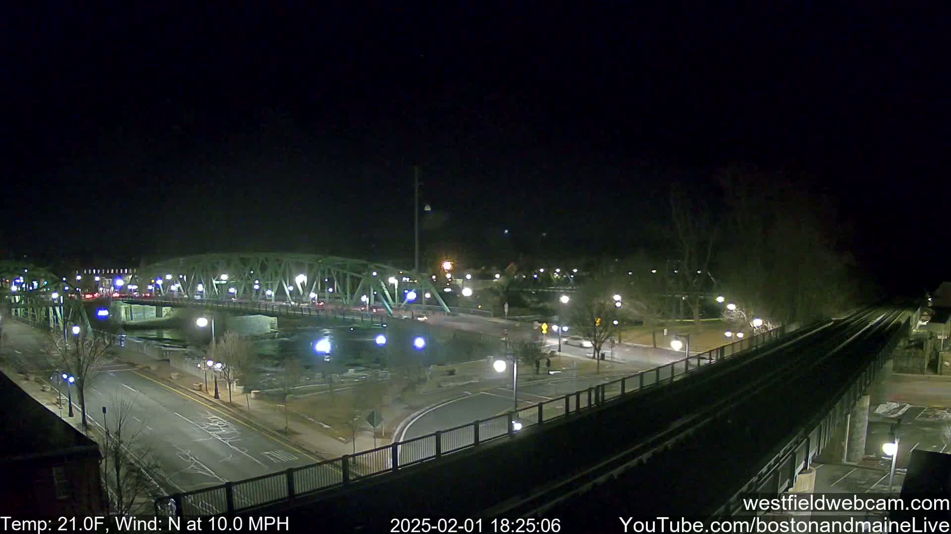 A nighttime view of a bridge over a river, with a road and train tracks intersecting, under a clear, cold sky with a north wind.