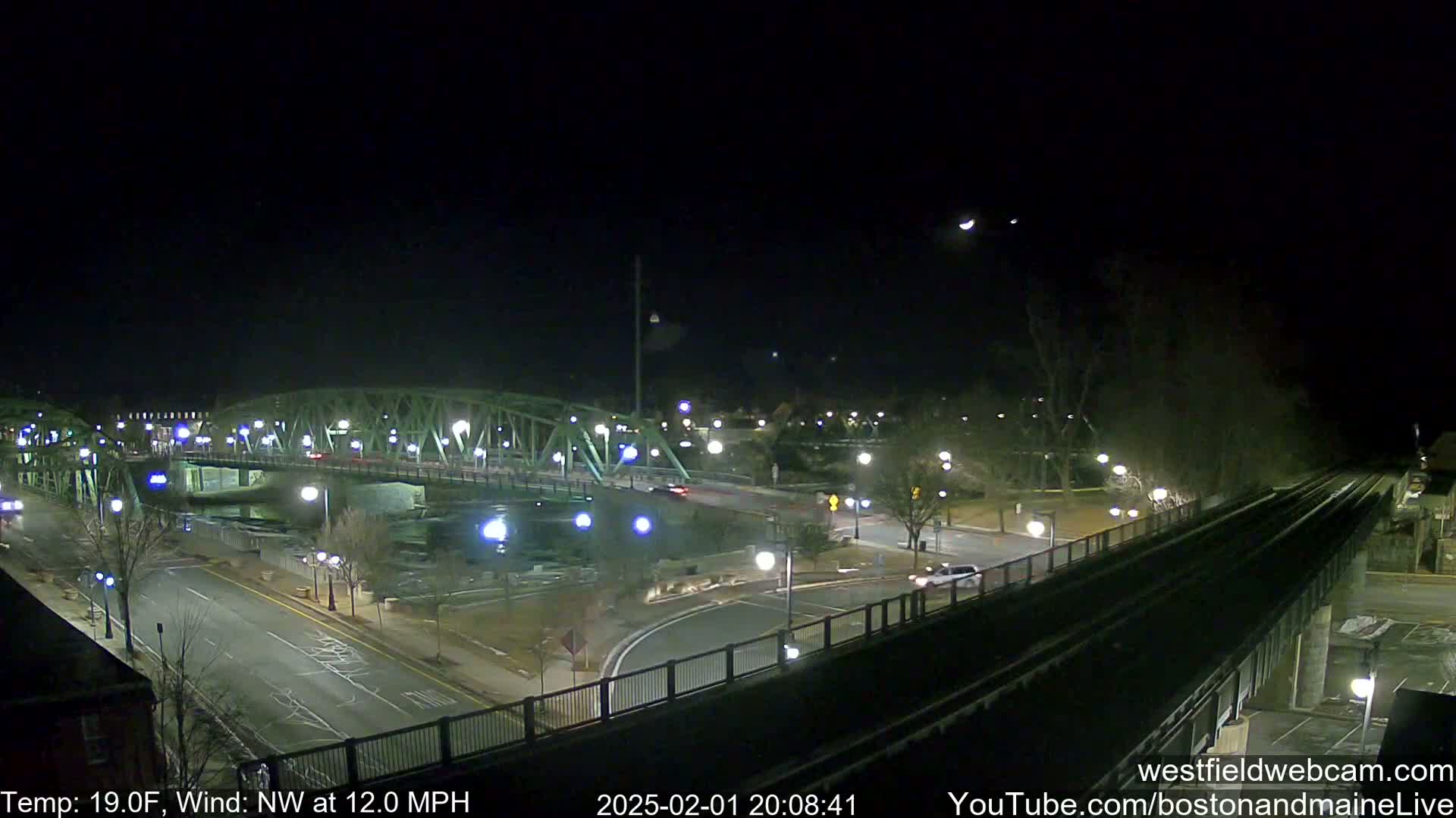 A nighttime view of a bridge over a river, with sparsely lit streets and a train track visible, under a clear, cold sky with a northwest wind.