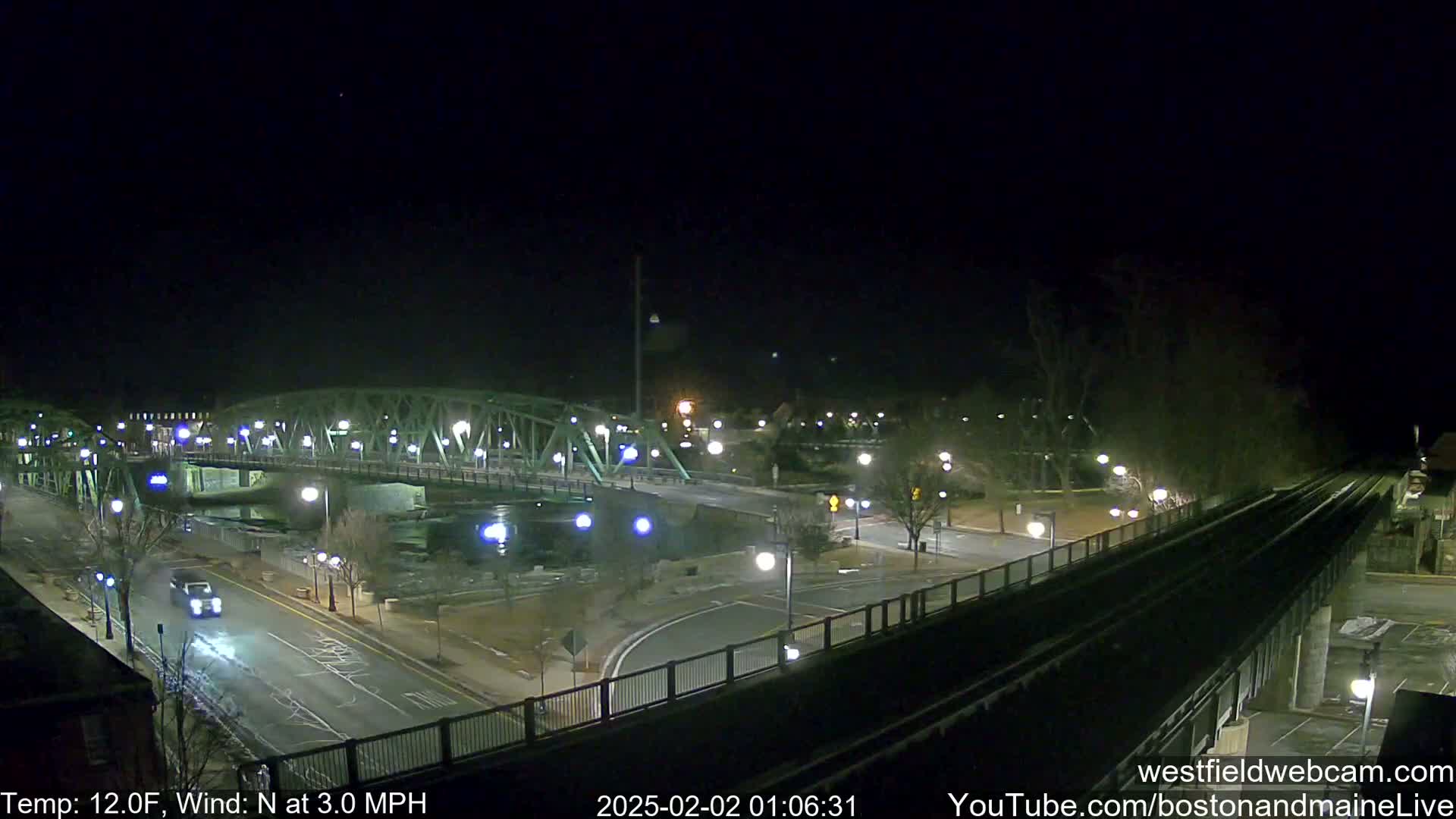 A nighttime view of a bridge over a river, with a road and train tracks intersecting below, under a clear, cold sky.