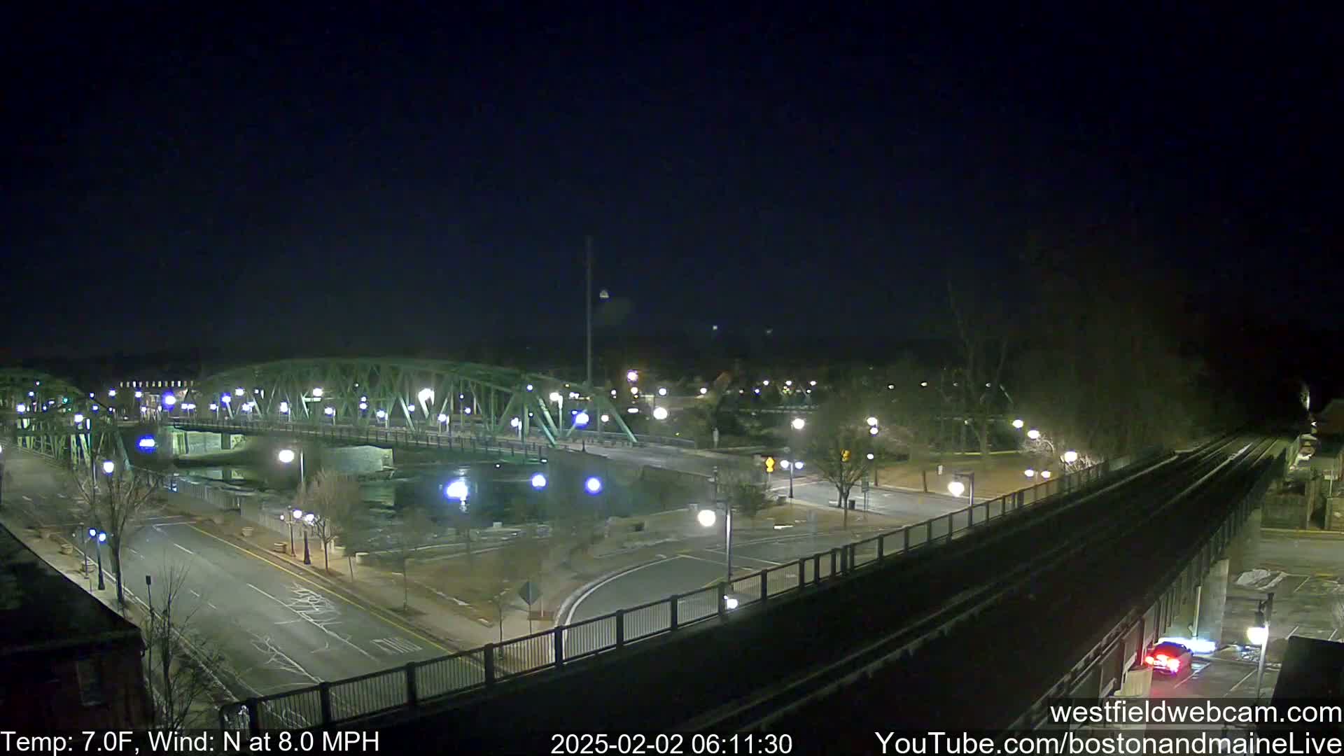 A nighttime view of a bridge over a river, with a road and train tracks intersecting nearby, under a clear, dark sky with a north wind at 8 mph and a temperature of 7°F.