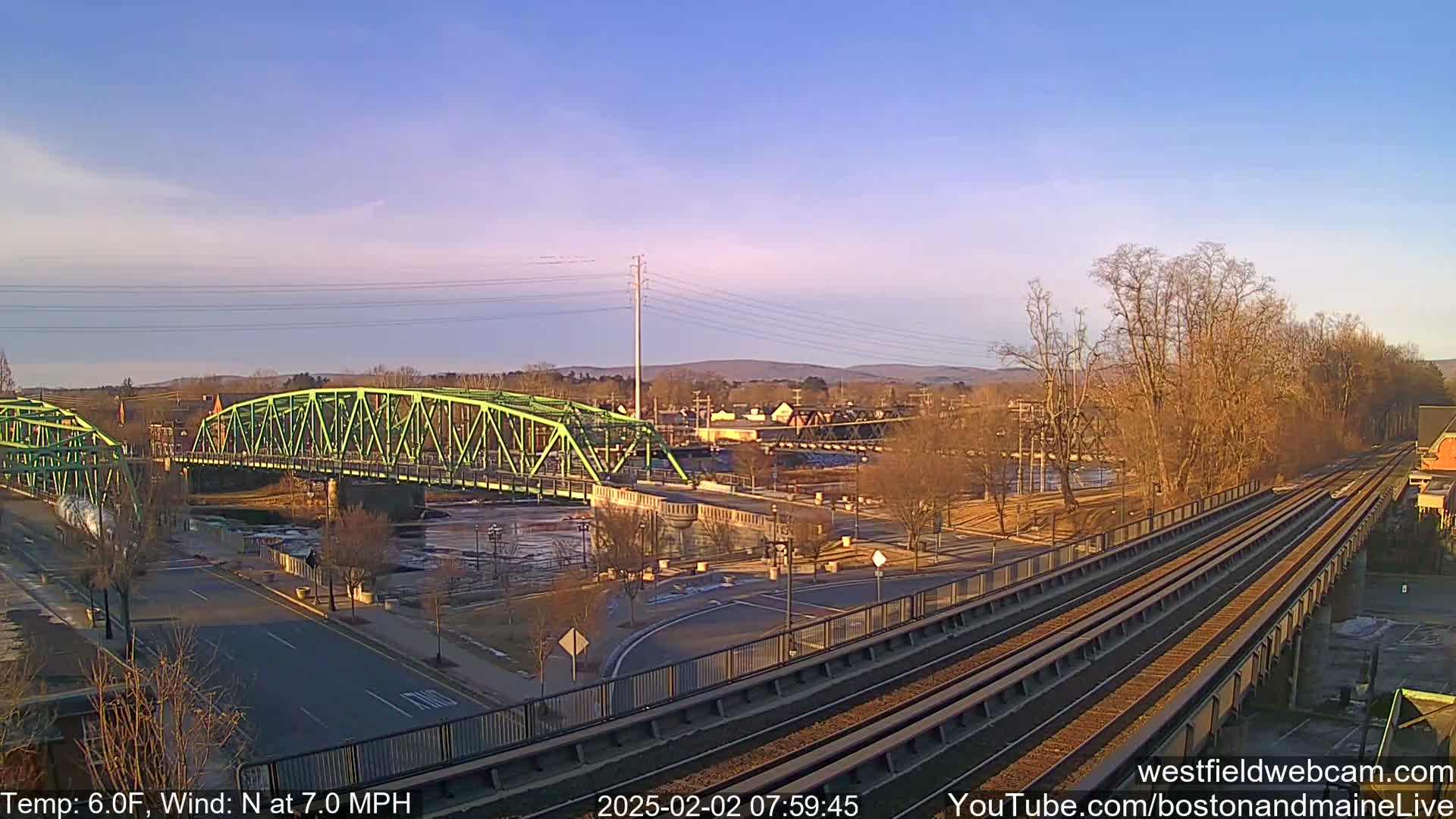 A bright, clear morning shows a lime-green bridge over a river, with train tracks in the foreground and bare trees lining the riverbank.