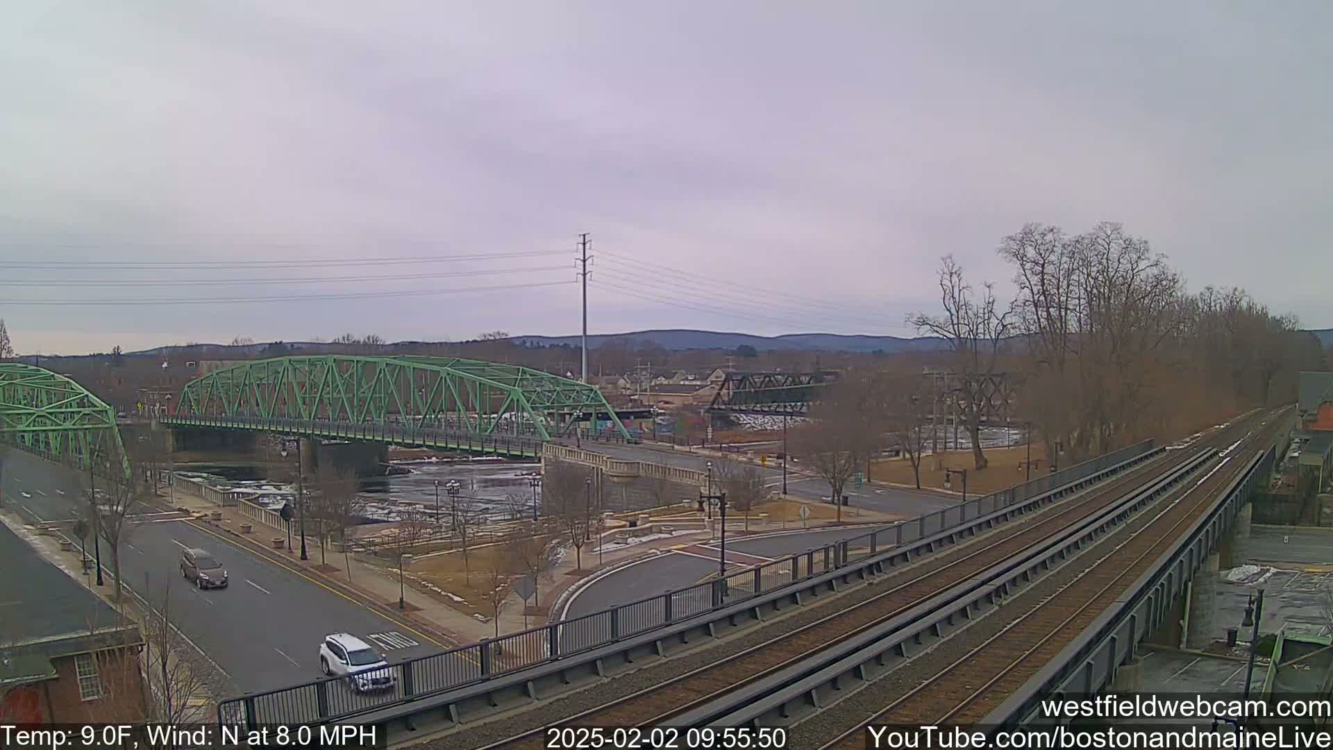 A green bridge spans a river with icy patches, alongside train tracks and a road, under an overcast sky with a temperature of 9°F and a north wind of 8.0 mph.