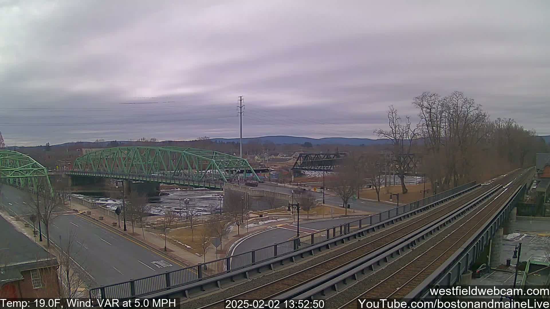 A view of two bridges over a partially frozen river, with train tracks and roadways leading towards them under an overcast sky, with a temperature of 19°F and a variable wind at 5.0 mph.