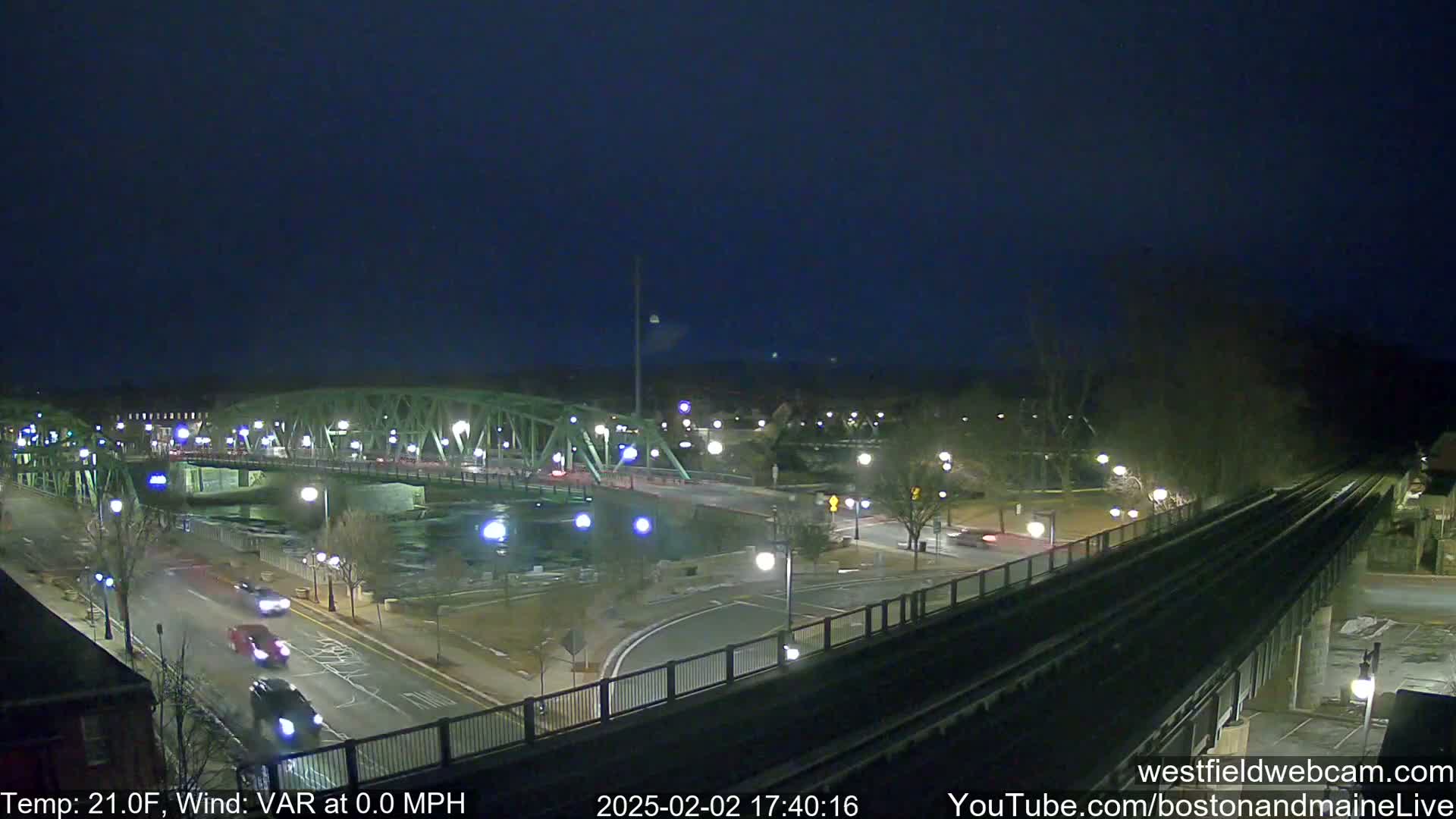 A nighttime view of a bridge over a river, with cars driving on a road beneath it and train tracks to the right, under a clear dark sky.
