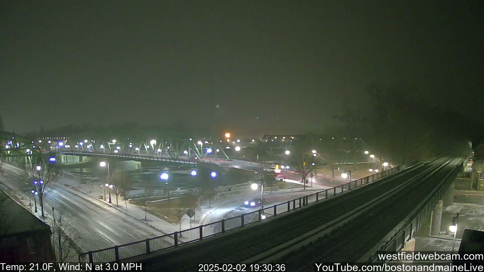 A lightly snow-covered bridge and road intersect near a river under a dark, nighttime sky.