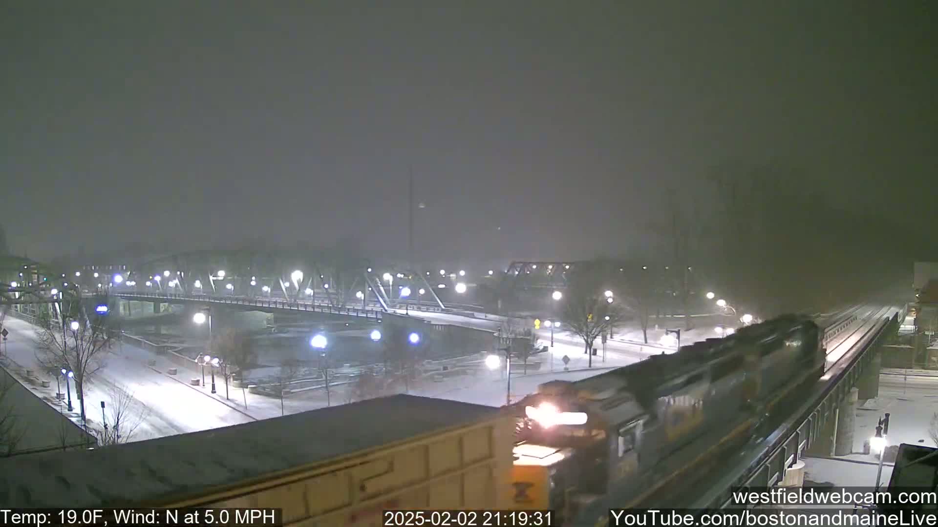 A train passes over a snow-covered bridge at night in a blizzard with a north wind of 5 mph and a temperature of 19 degrees Fahrenheit.