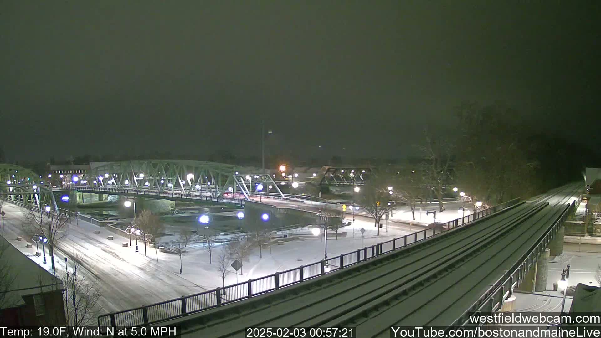 A snow-covered nighttime scene shows a bridge over a river, a train track, and a park with streetlights, under a dark sky.