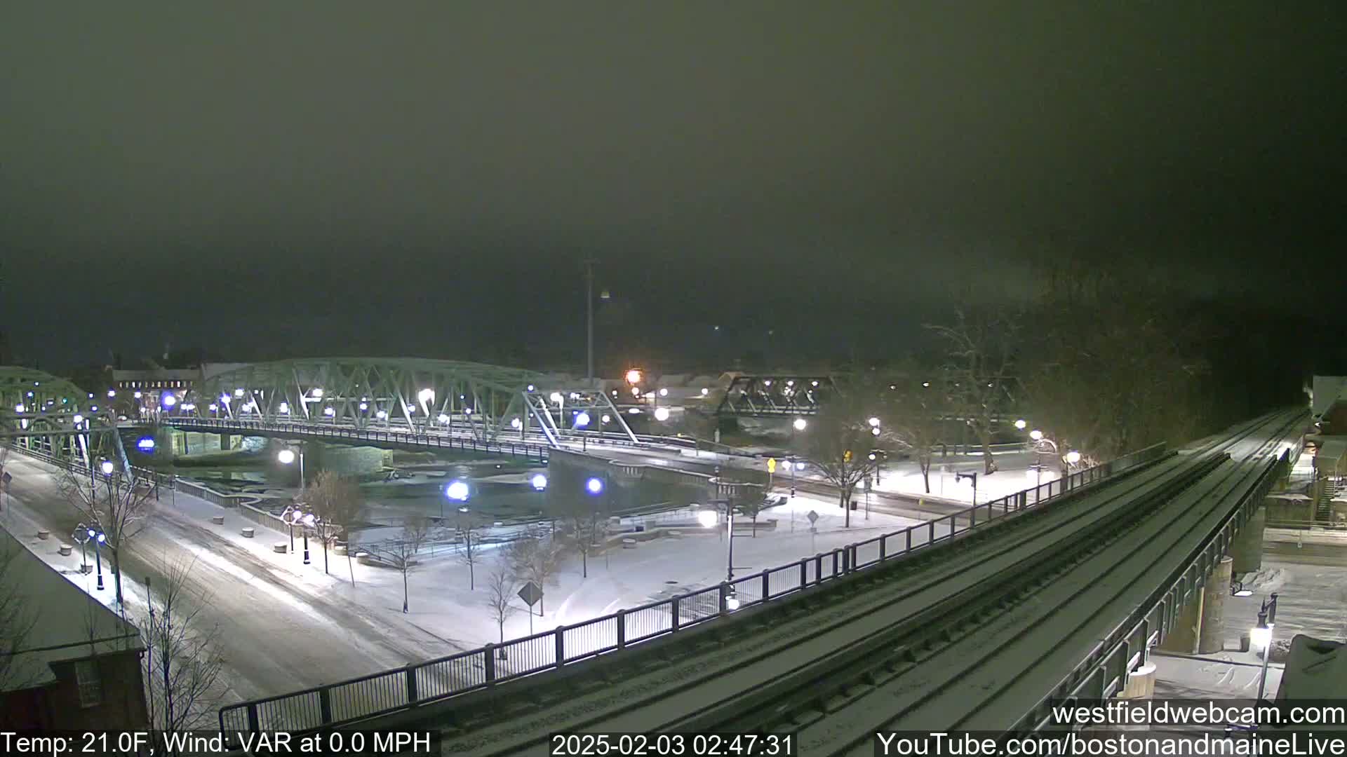 A snow-covered bridge and roadway at night, with train tracks in the foreground under a dark, overcast sky.