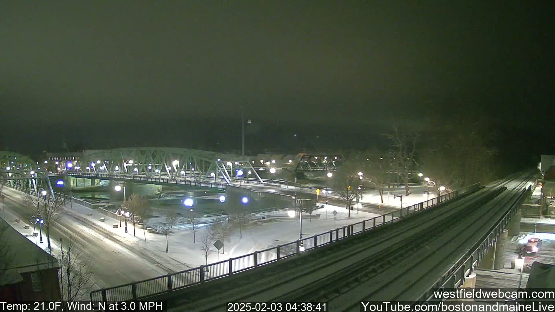 A snow-covered nighttime scene shows a bridge over a river, with train tracks and a road alongside, under a dark, hazy sky.