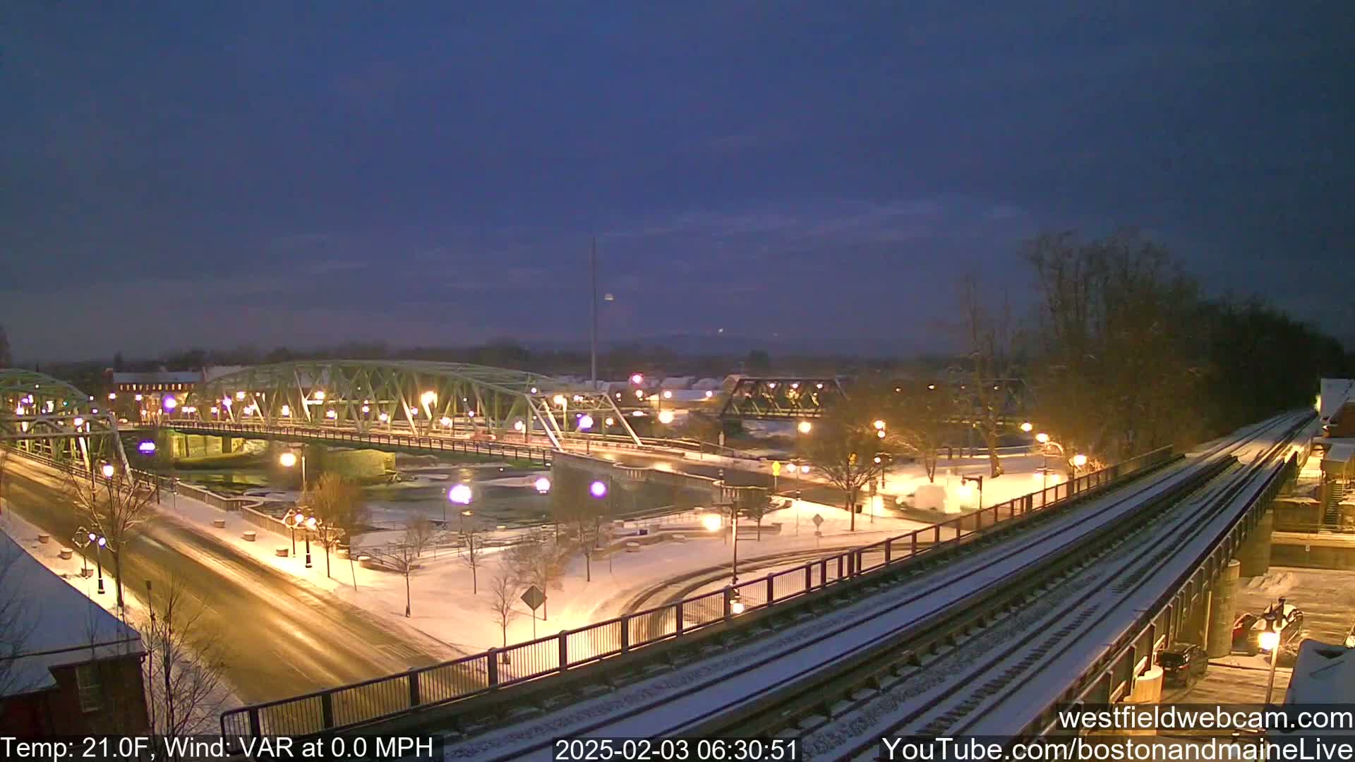 A snow-covered cityscape at night, featuring two bridges over a river, illuminated streets, and train tracks.