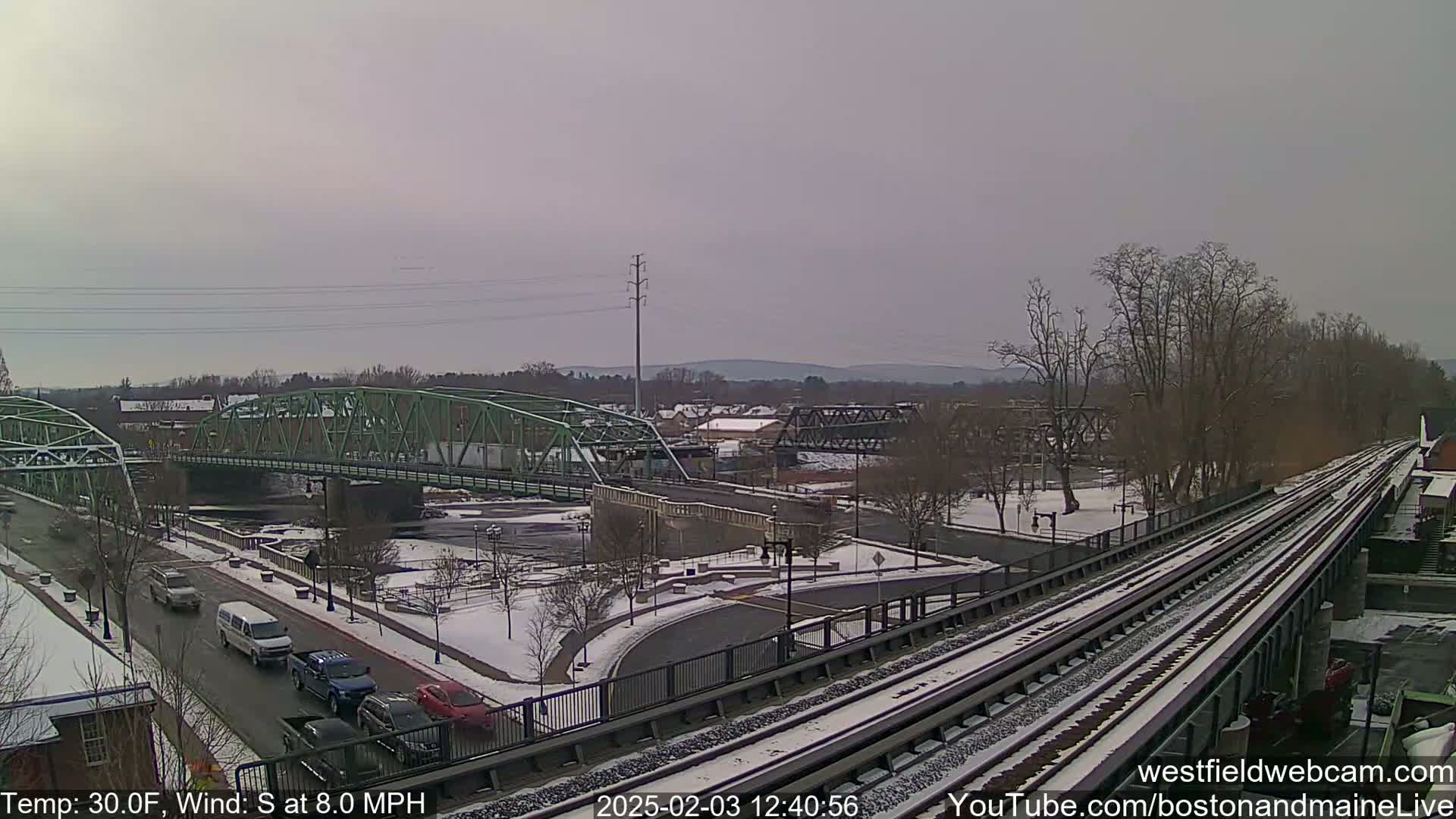 A view of two bridges over a river, with light snow on the ground and some cars on the roads, under an overcast sky.
